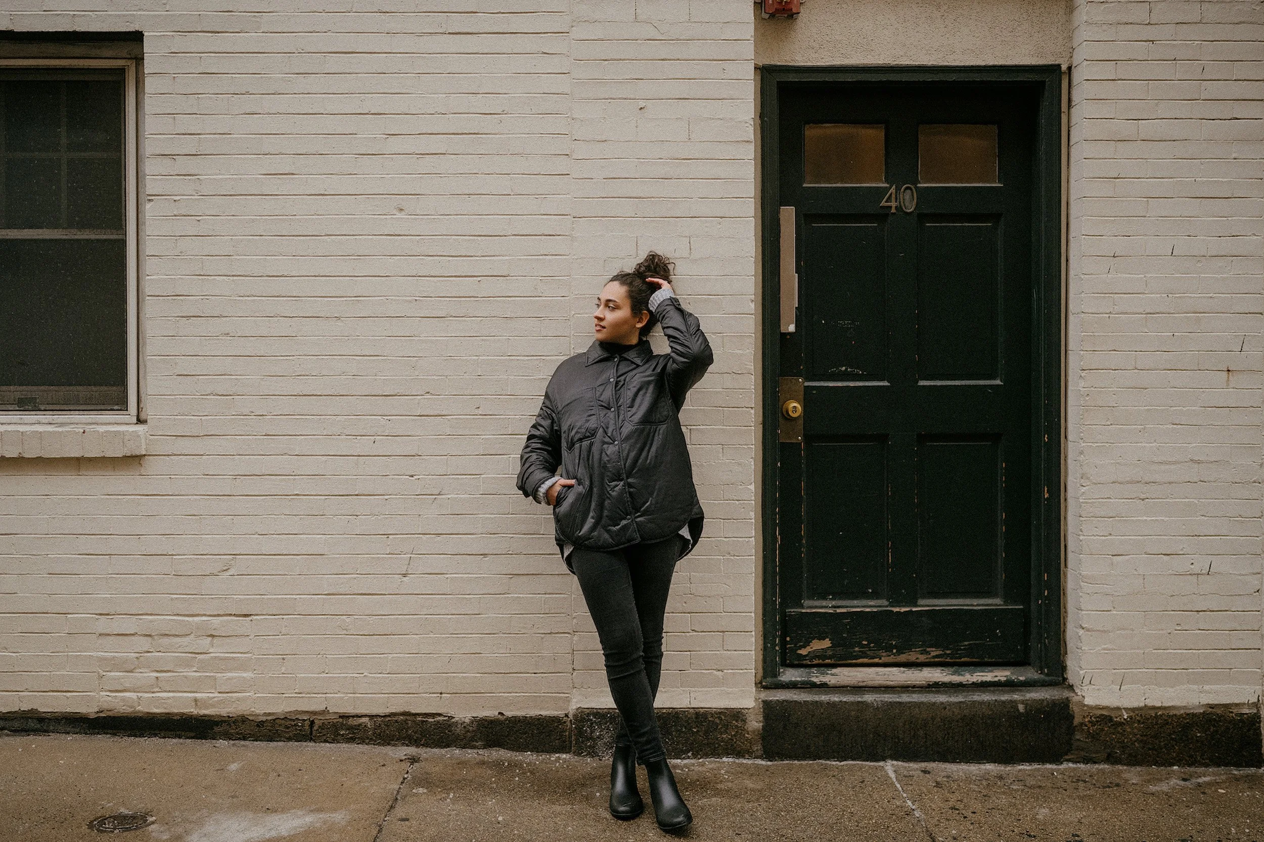 A woman standing outside a building with a black door numbered 40, posing with her hand on her head, wearing a black jacket, black pants, and black boots, on a rainy day.