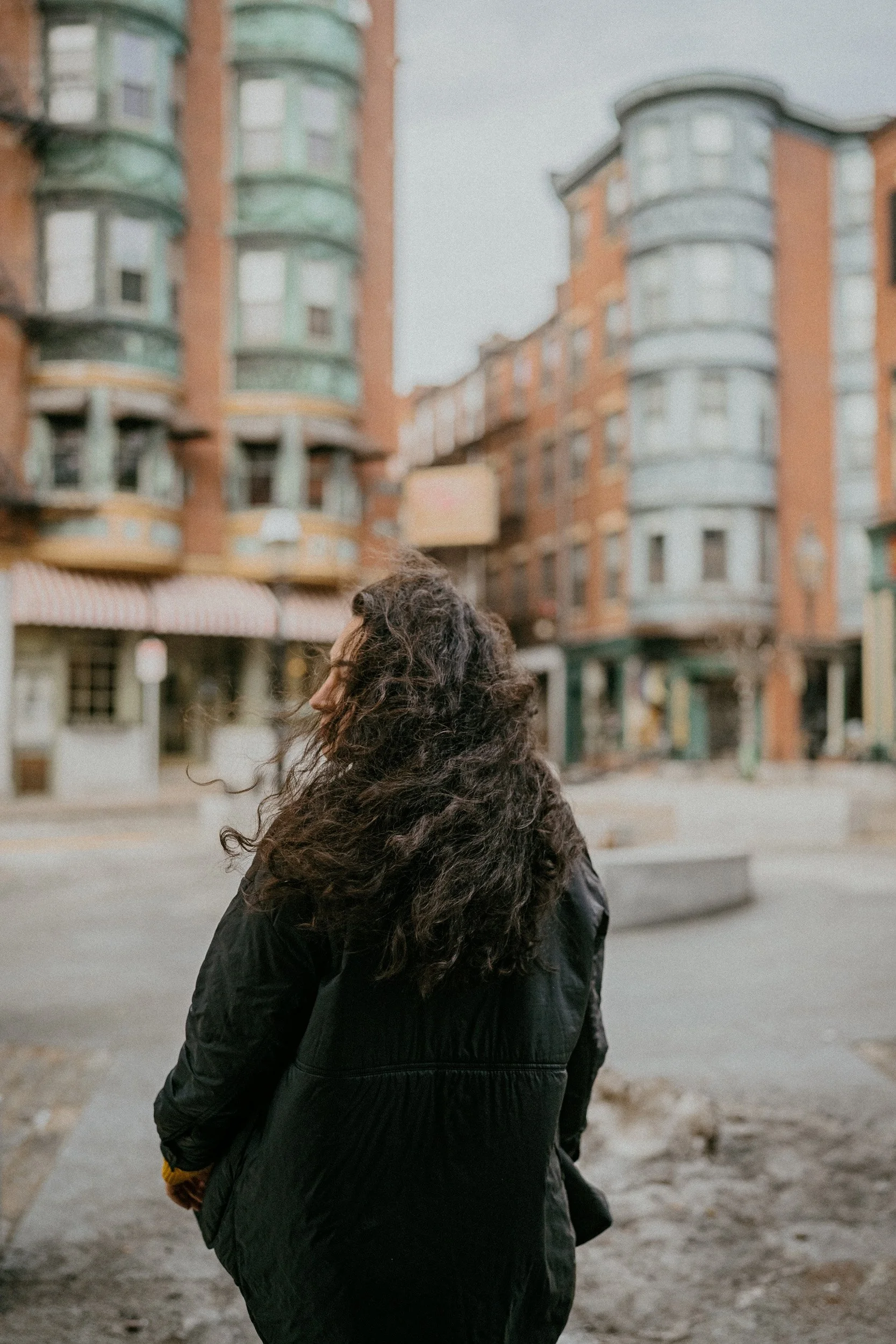 A woman with long curly hair wearing a black jacket, standing outdoors with her back toward the camera, in front of colorful apartment buildings.