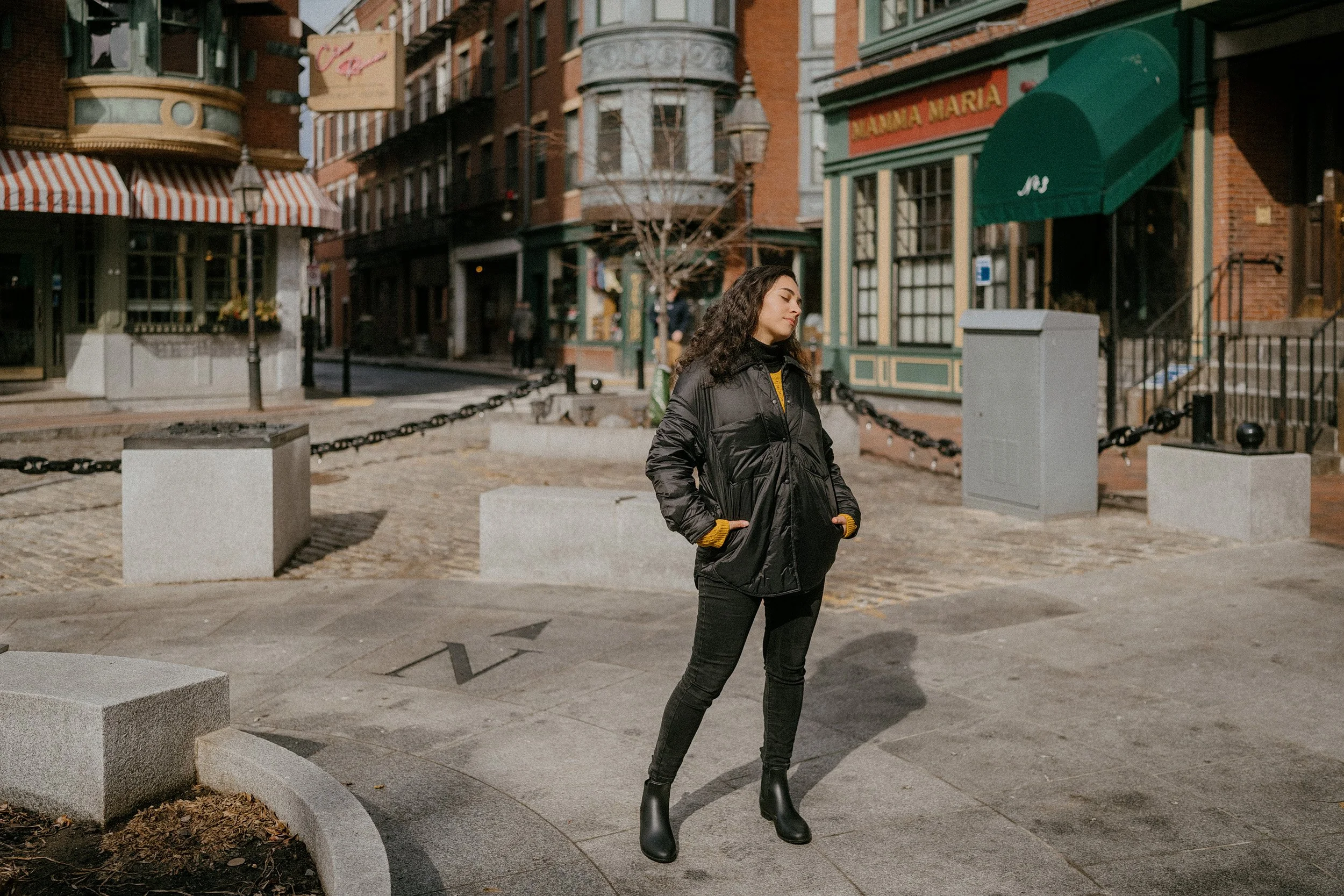 A woman with curly dark hair standing on a city sidewalk in front of colorful storefronts and buildings, wearing a black puffer jacket, black pants, and black ankle boots, with her hands in her pockets and eyes closed.