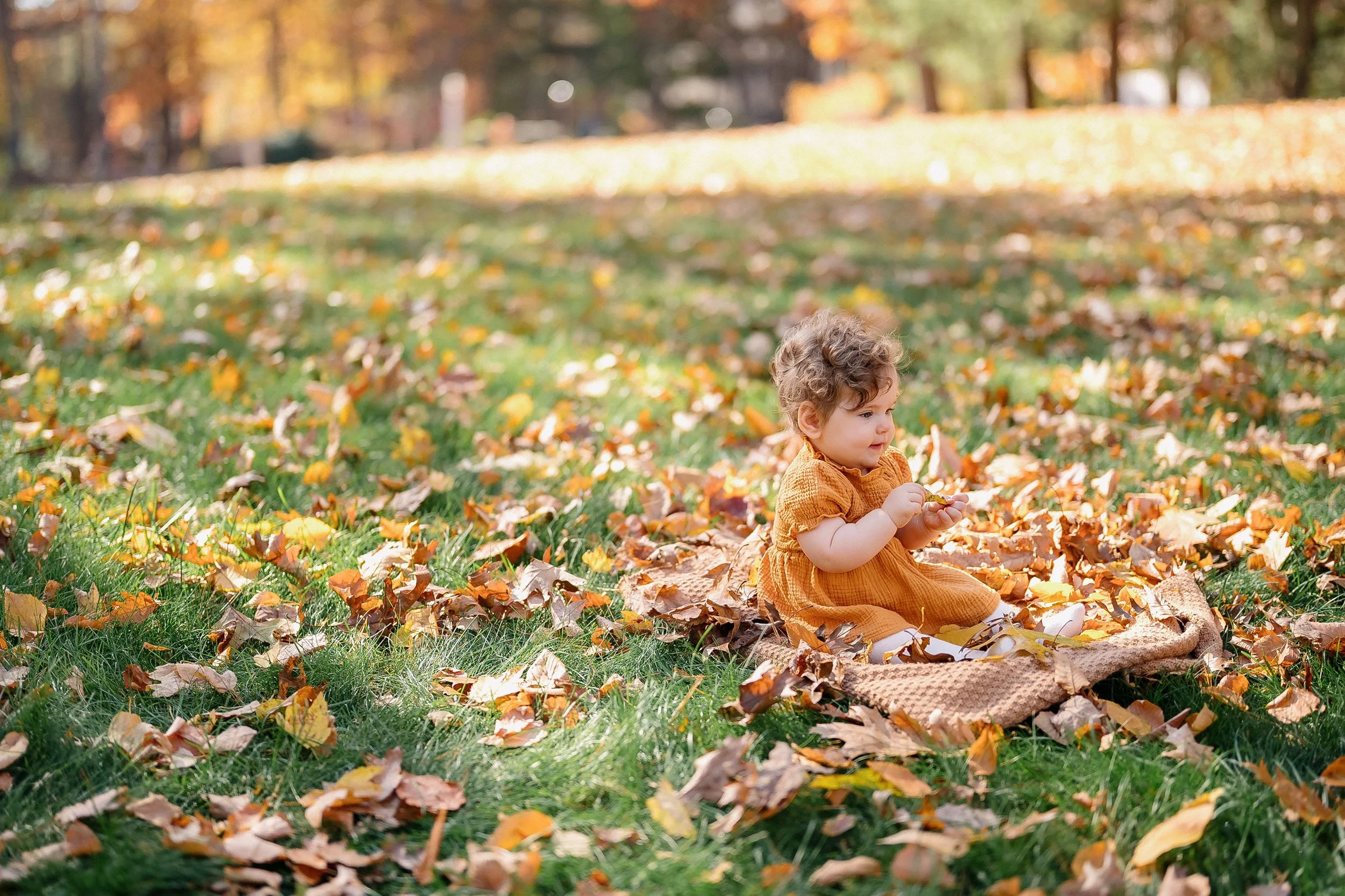 A young girl sitting outdoors on a blanket in a park, surrounded by fallen autumn leaves, holding a leaf in her hands.