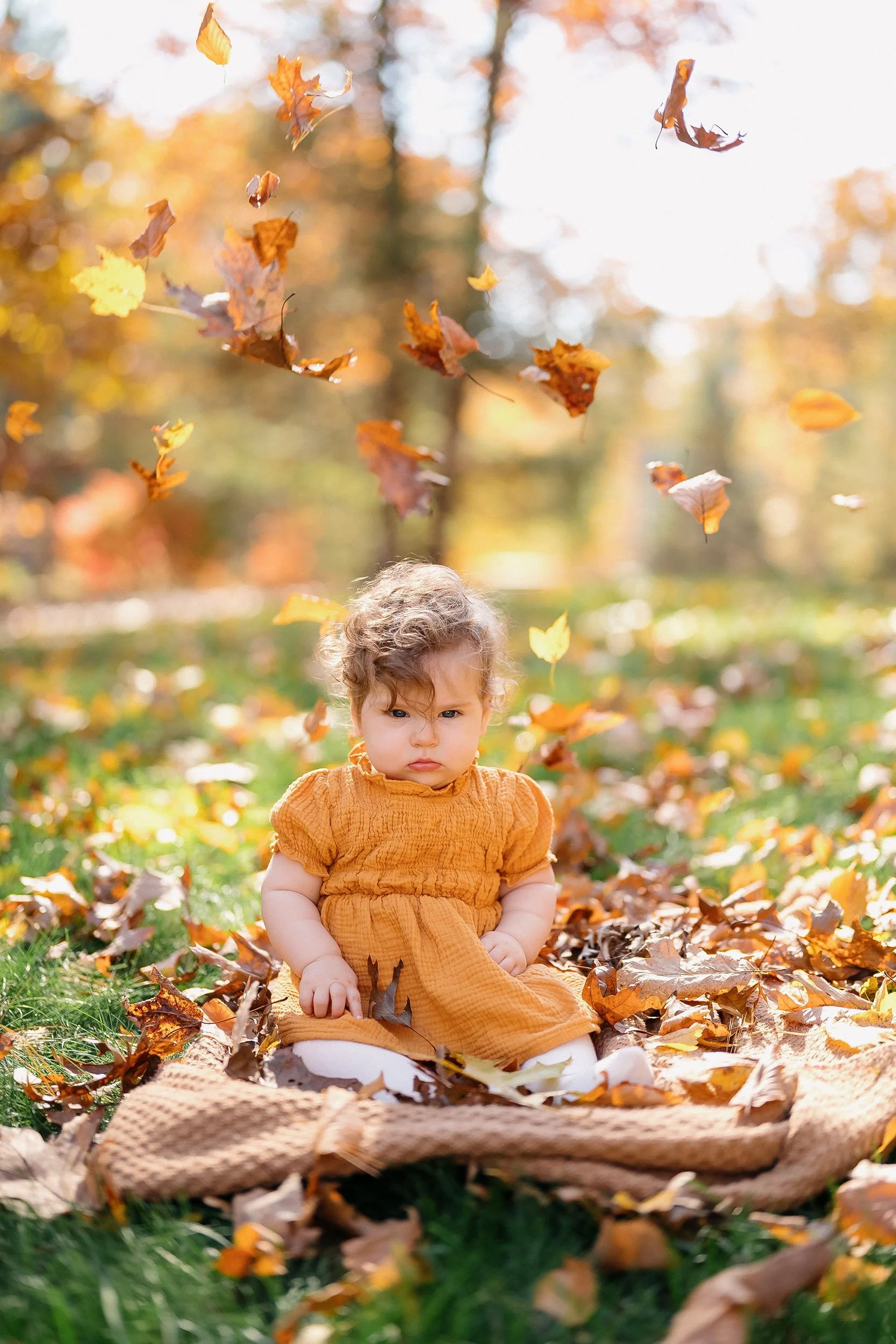 A young girl with curly hair sitting outdoors on a blanket among fallen autumn leaves, with leaves falling around her on a sunny day.