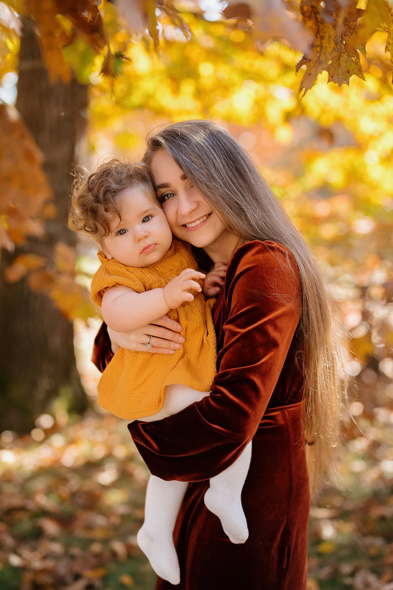 A young woman smiling and holding a toddler in her arms outdoors surrounded by autumn leaves and trees.
