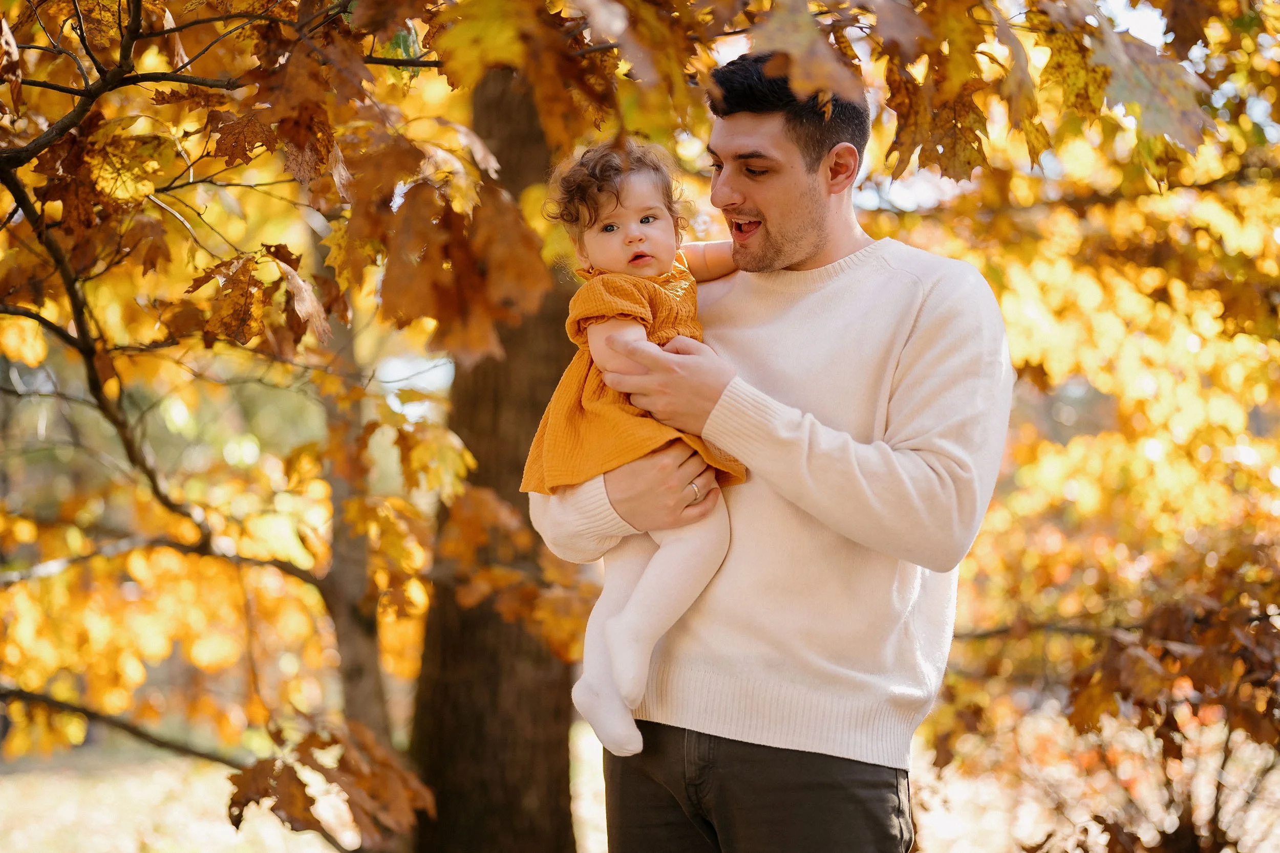 A man holding a young girl in an autumnal outdoor setting with trees and orange leaves.