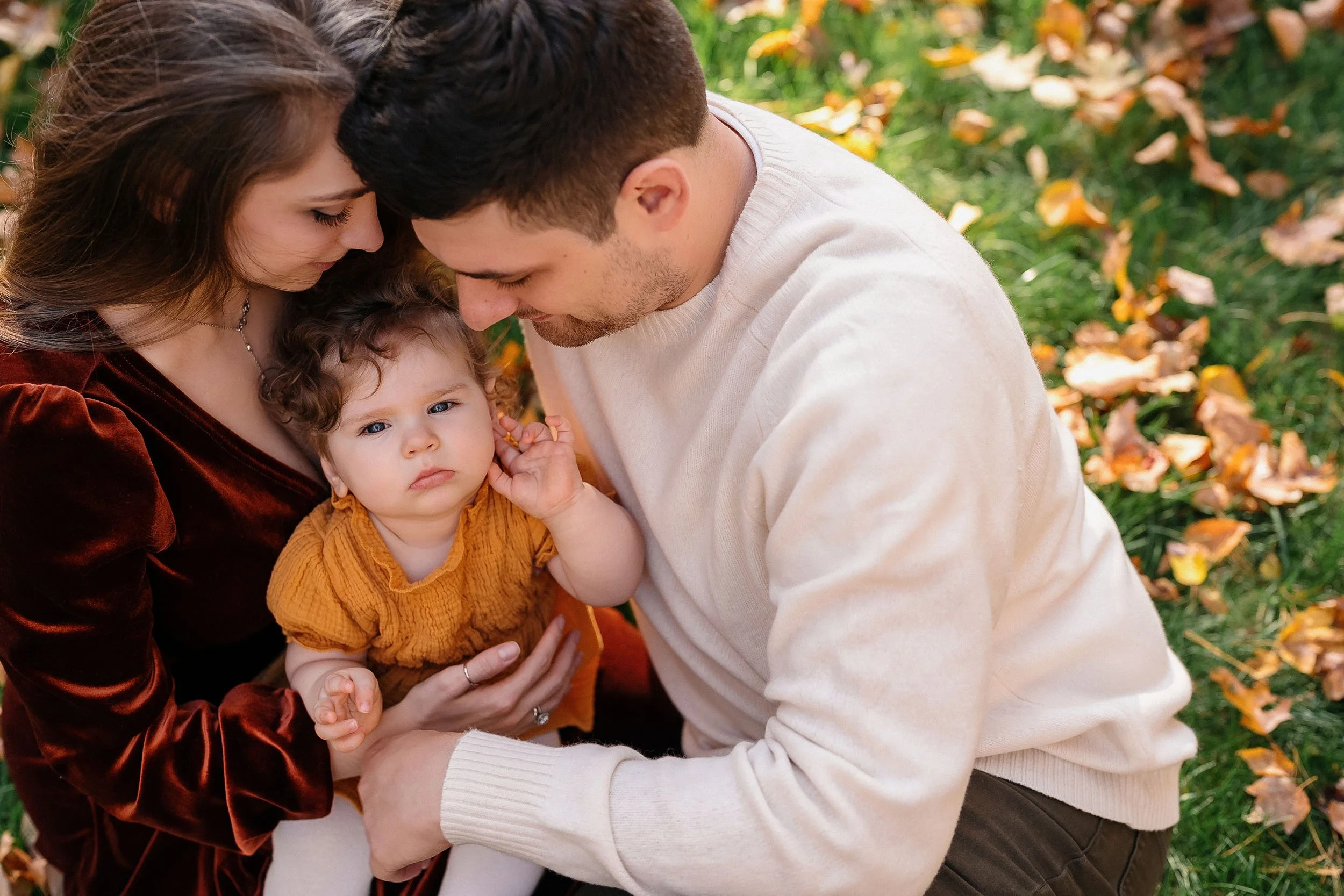 A family of three sitting on grass with fallen autumn leaves, looking affectionately at their young daughter.