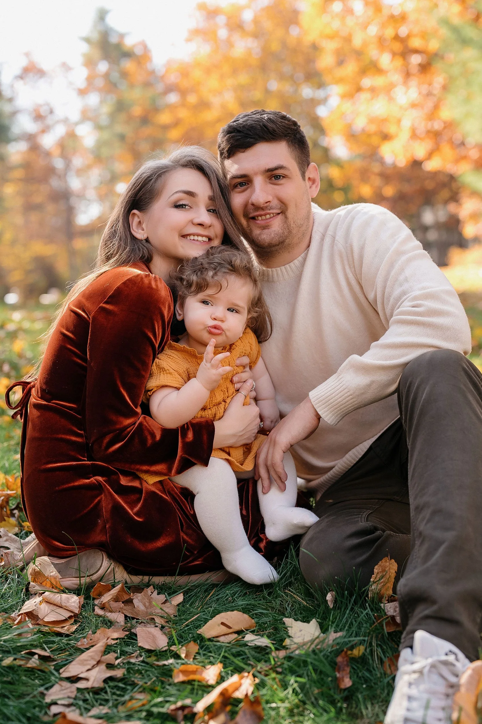 A family of three sitting outdoors in autumn, with colorful fall trees in the background. The woman is wearing a velvet dress, the man is in a beige sweater, and the little girl is making a duck face, wearing a mustard dress and white tights.