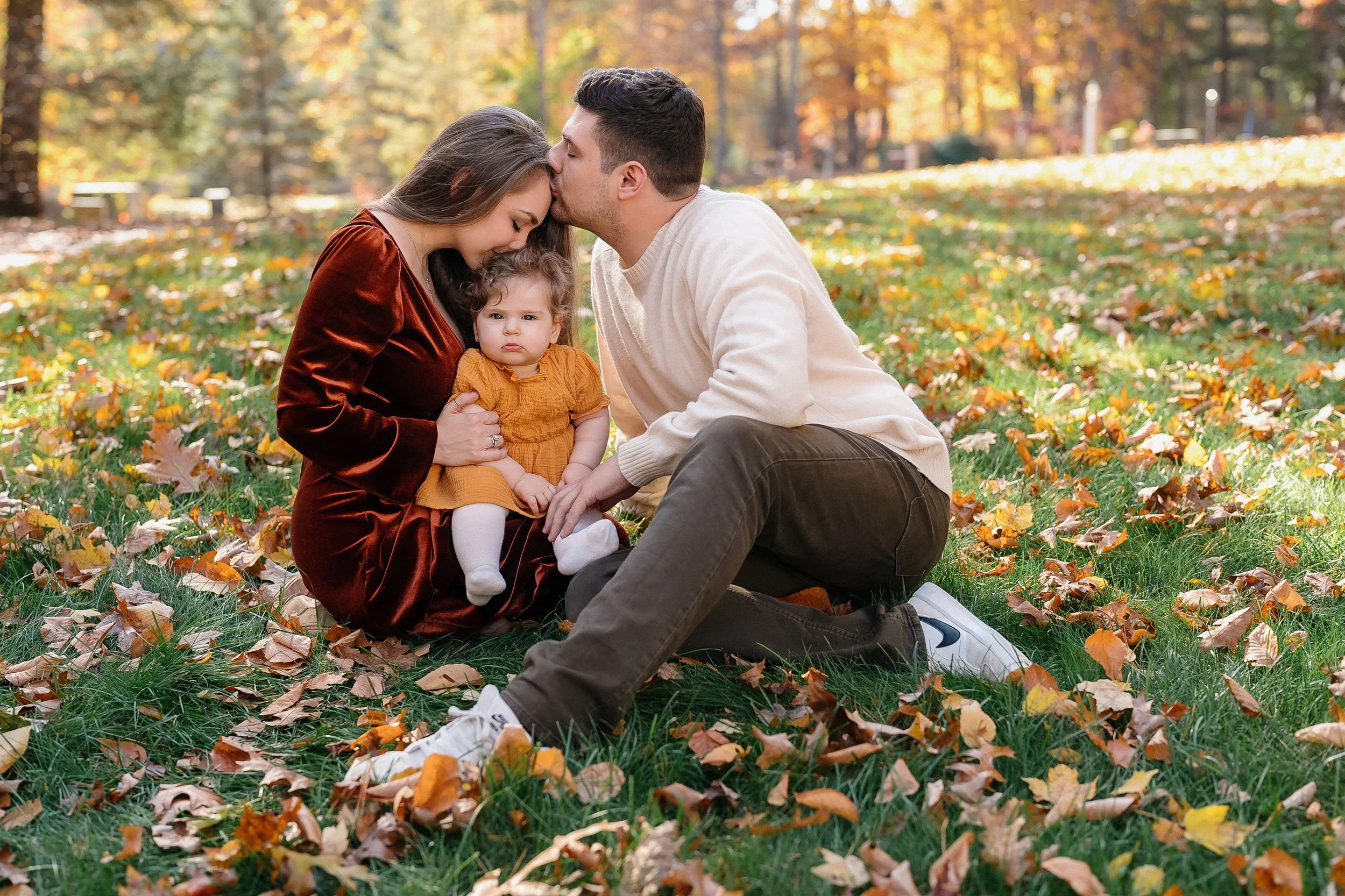 A family of three sitting on grass with autumn leaves, the father and mother kissing the child's forehead.