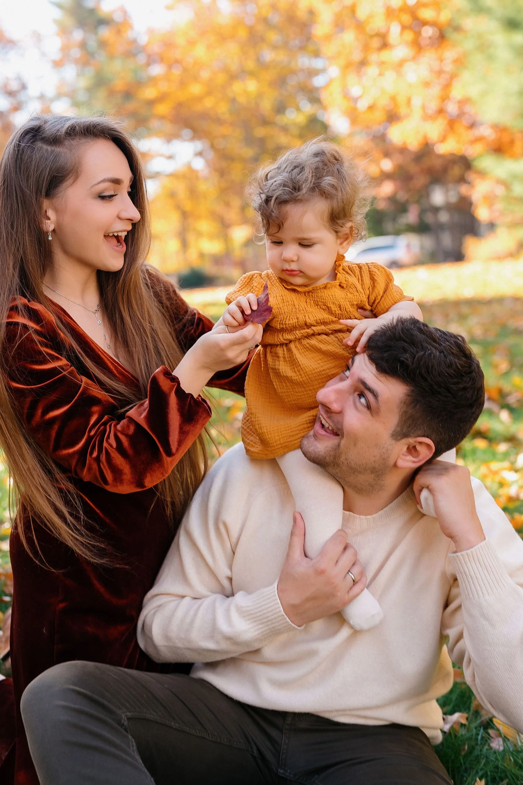 Family enjoying an autumn day outdoors with colorful fall leaves, a woman holding a leaf, a man lifting a young girl onto his shoulders.