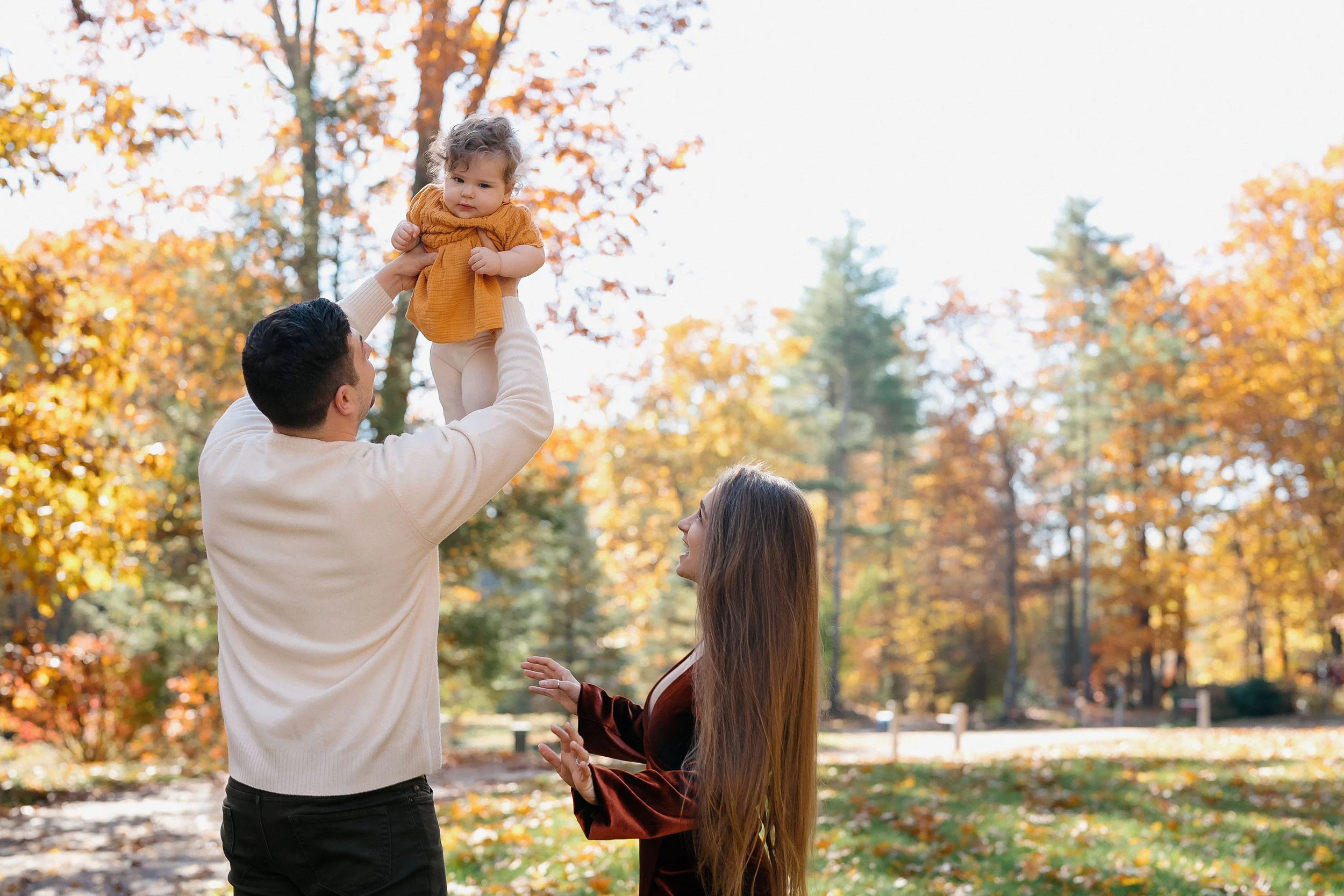 A family of three enjoying the outdoors in a park during autumn; the father lifts a young girl with curly hair and a yellow sweater, while the mother reaches out toward them surrounded by trees with colorful fall foliage.