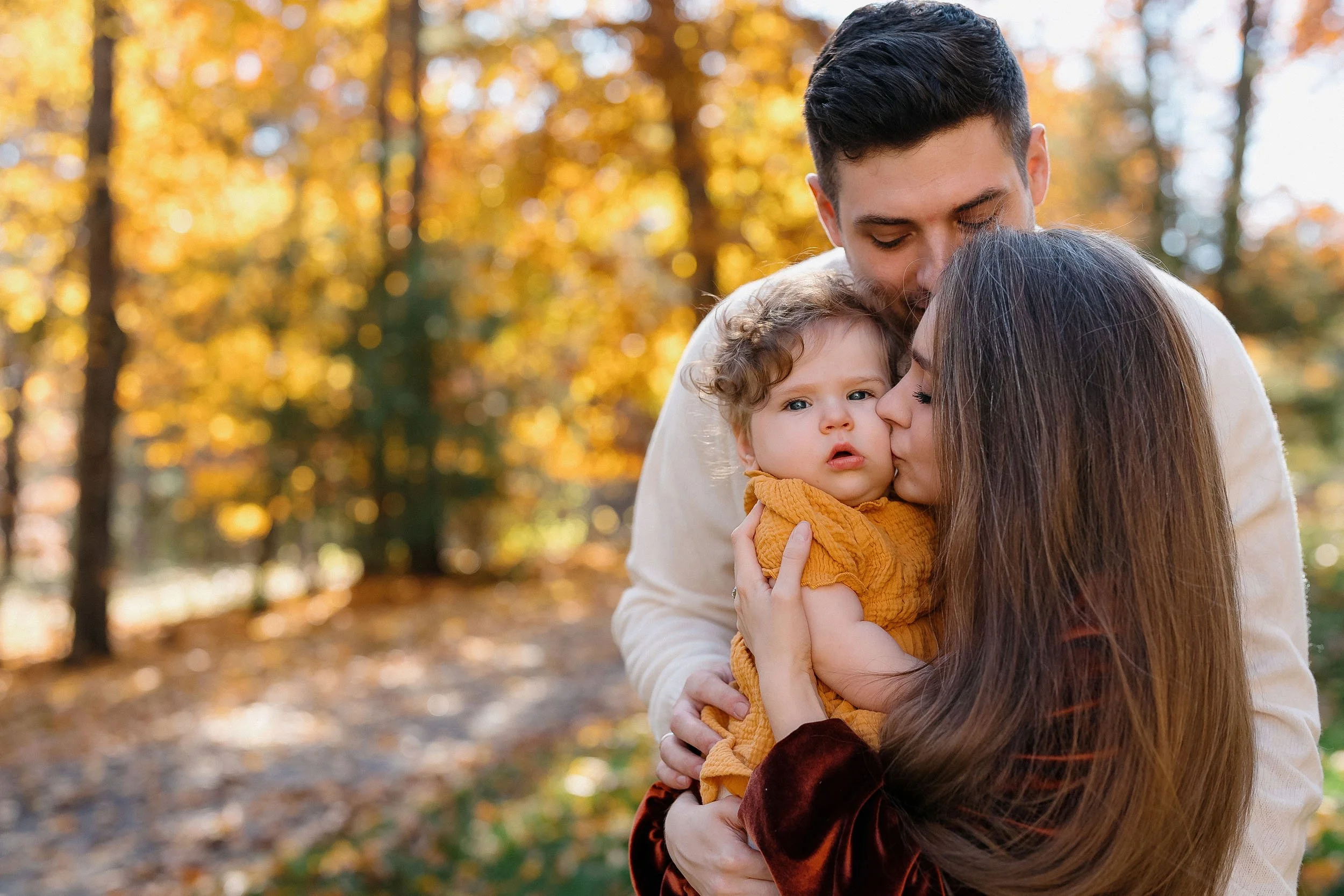 A family of three hugging outdoors in a park with autumn trees in the background, the mother kissing the child on the cheek and the father holding them.