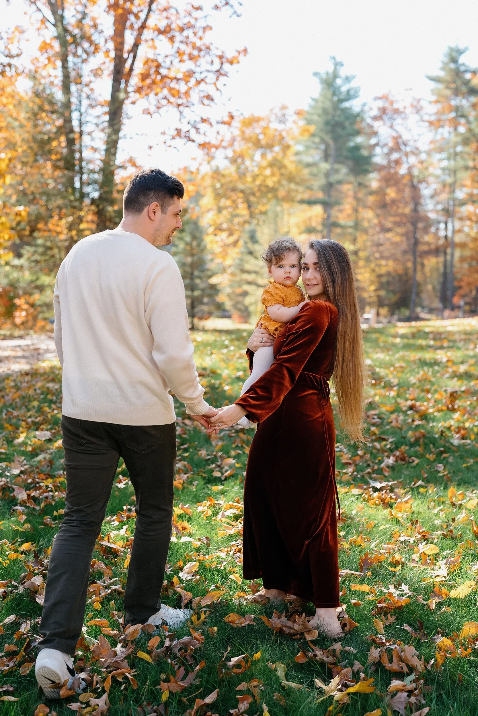 A family of three holding hands outdoors in an autumn park, with colorful fall leaves on the ground and trees in the background.