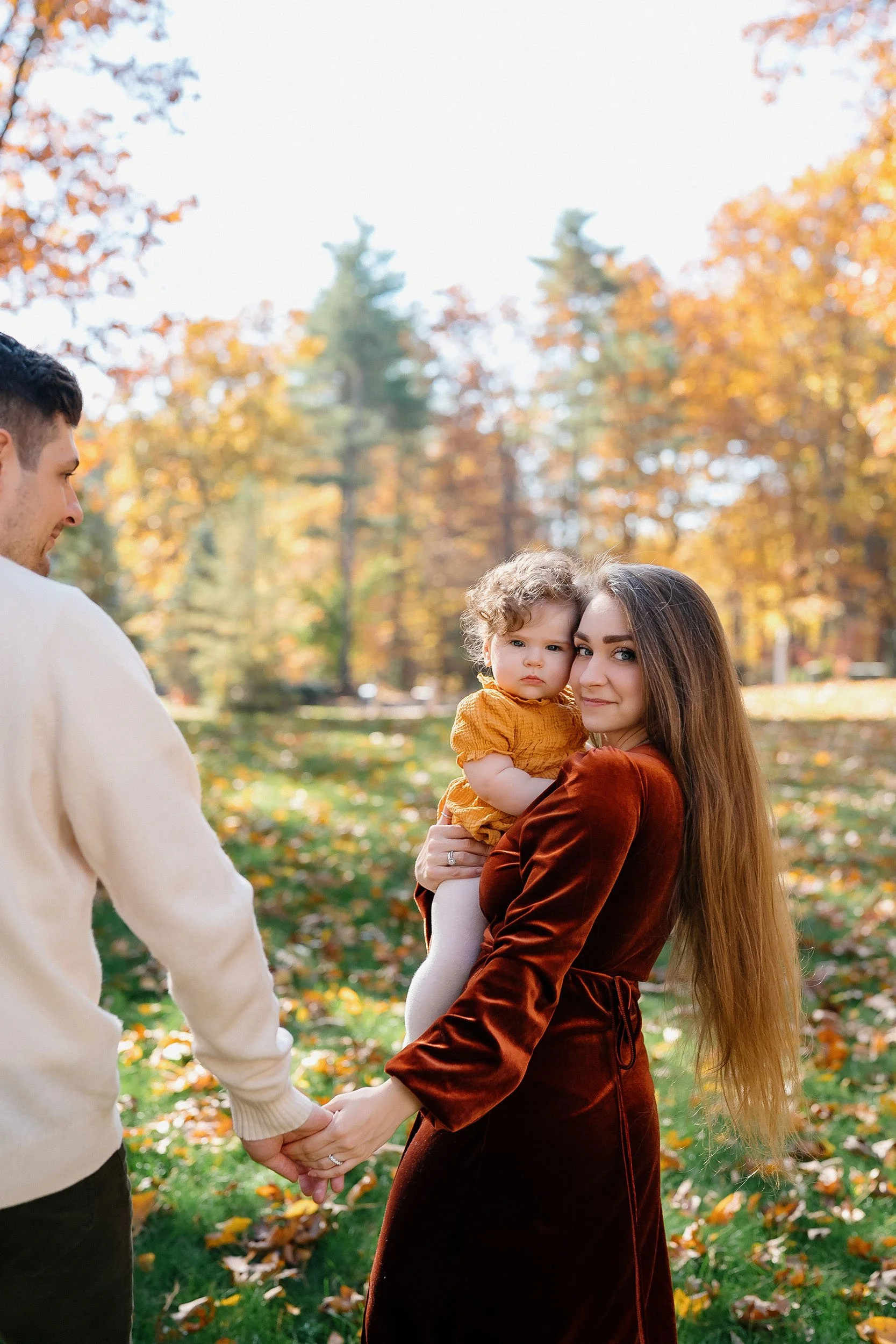 A woman holding a young child outdoors in a park with fall foliage, holding hands with a man.
