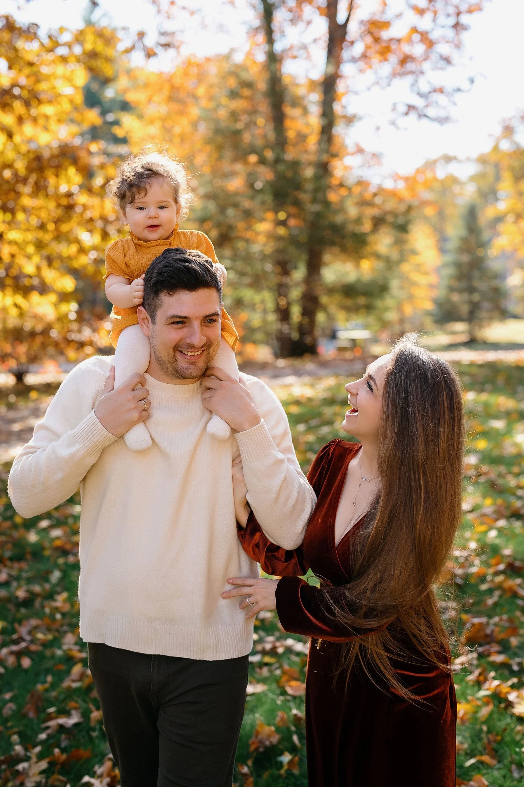 A family of three smiling outdoors in a park with autumn leaves. The father carries a young girl on his shoulders, and the mother stands beside them.