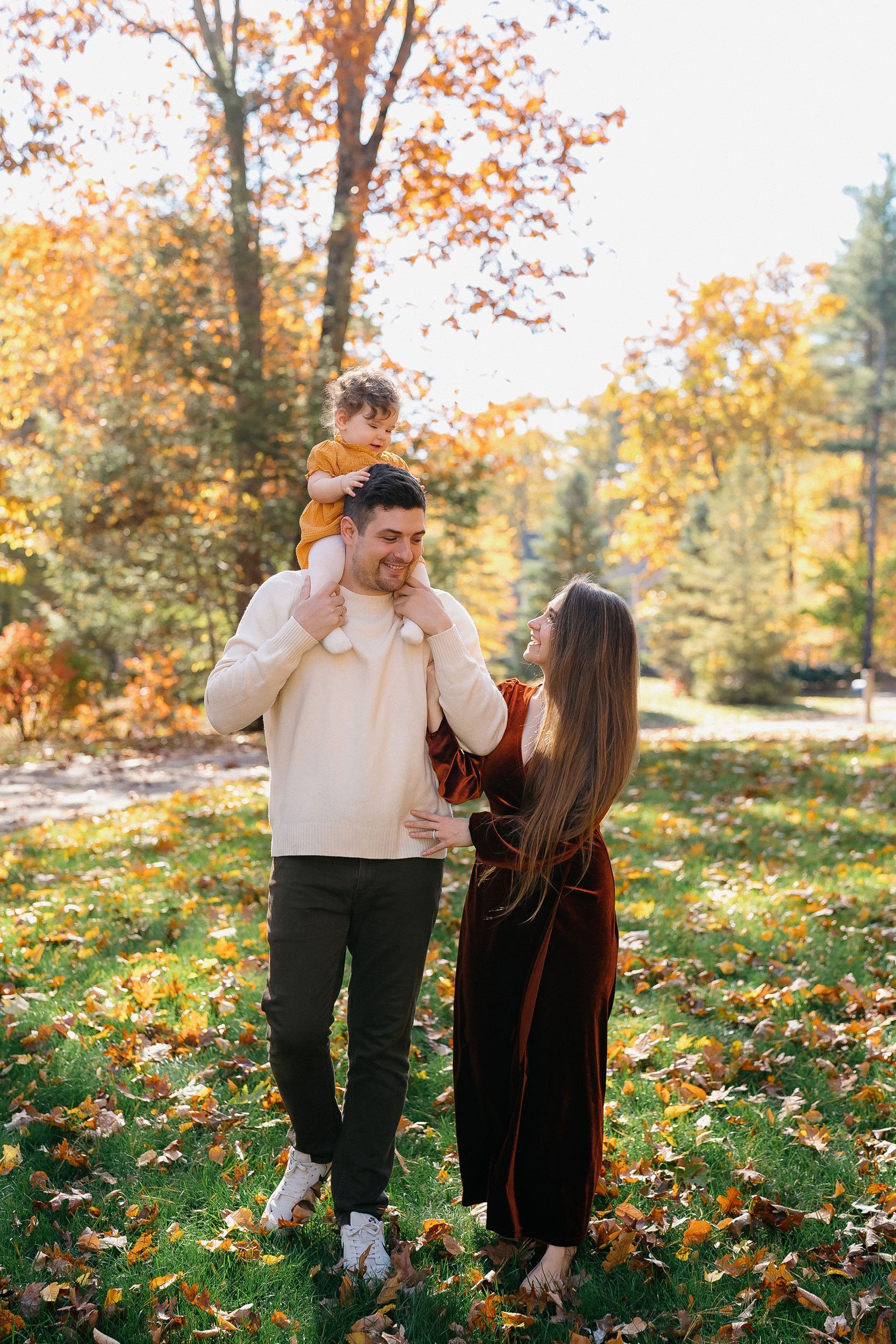 A family of three enjoying fall in a park, with colorful autumn leaves on the ground and trees in the background. The father carries a young girl on his shoulders, and the mother is smiling at them.