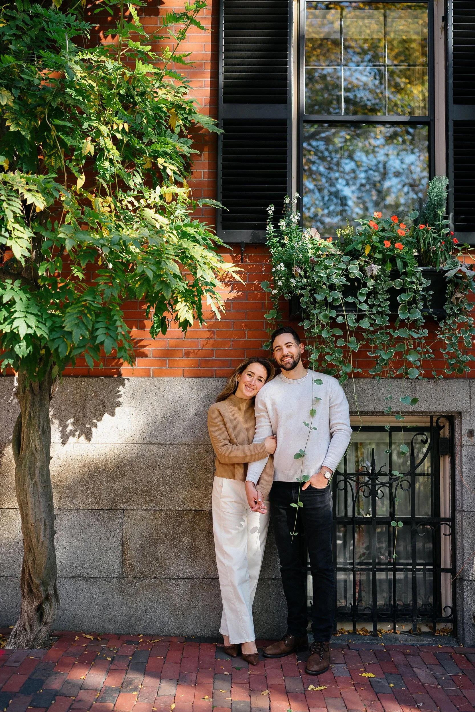 A smiling man and woman standing close together outdoors in front of a brick building with a window and black shutters, surrounded by green plants and trees, holding hands and leaning on each other.