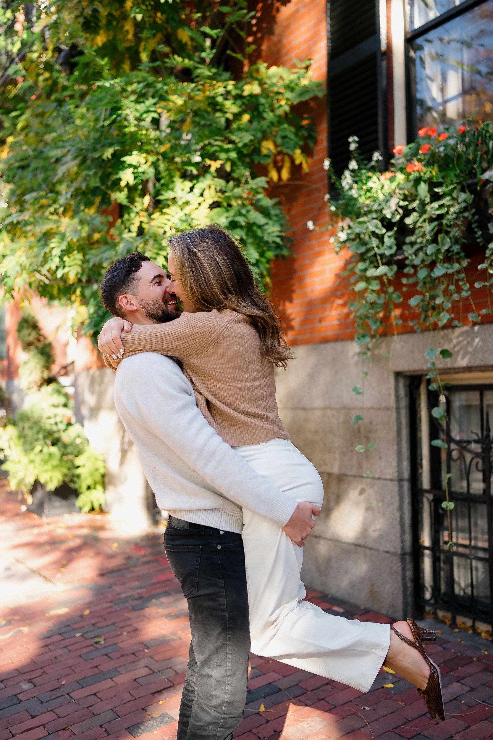 A man lifting a woman in an embrace on a brick sidewalk outdoors with greenery and a brick building in the background.