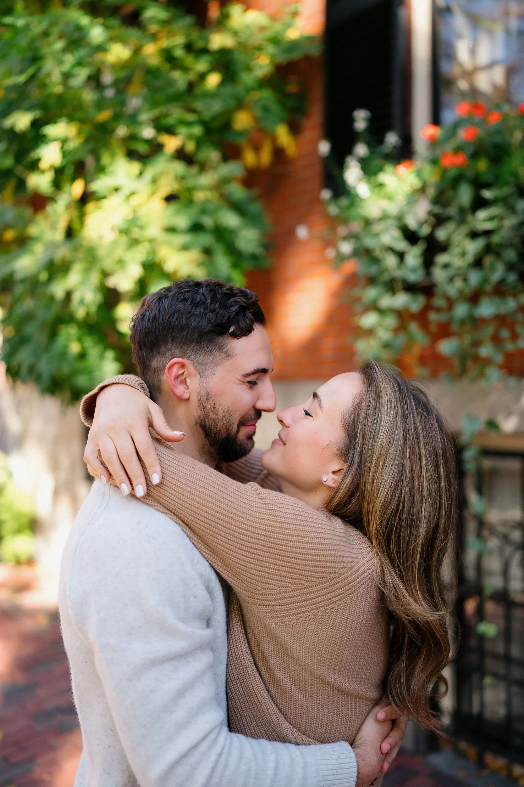 A couple embraces and looks into each other's eyes outdoors on a sunny day, with trees and a brick building in the background.