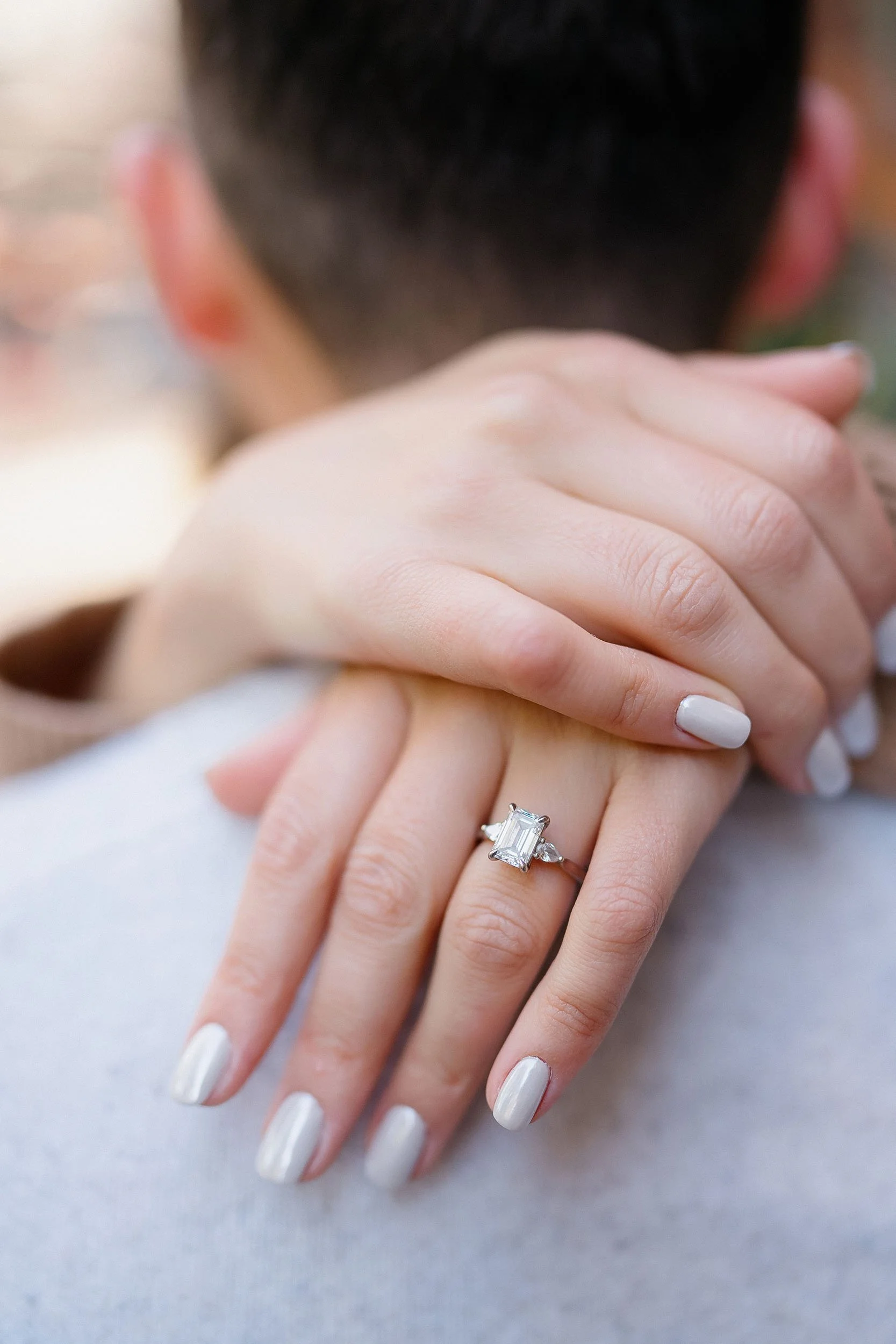 A close-up of a woman's hand with a large emerald-cut diamond engagement ring, resting on another person's hand with a background of blurred outdoor scenery.