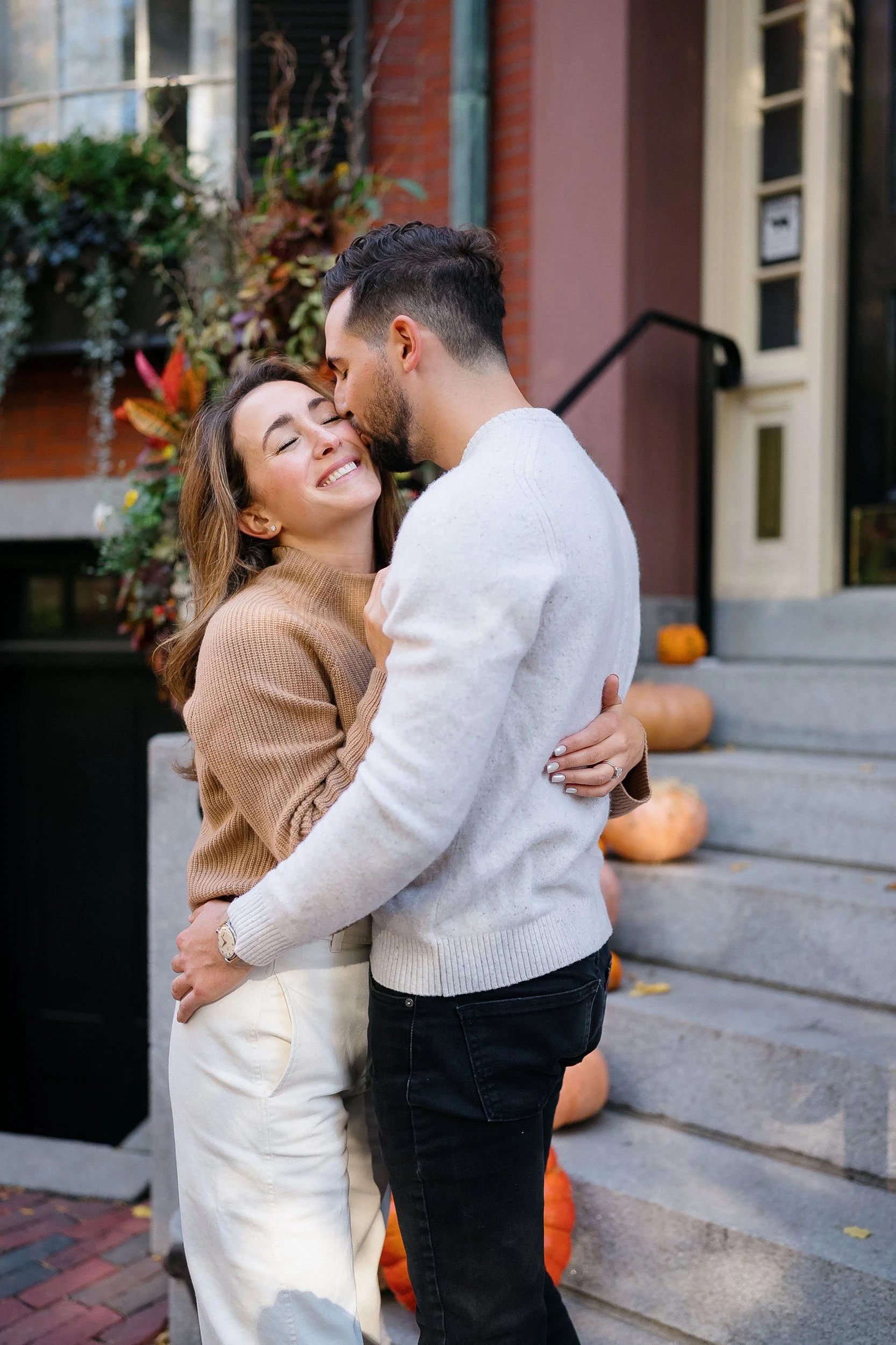 A couple hugging and smiling outside on a fall day in front of stairs decorated with pumpkins and foliage.
