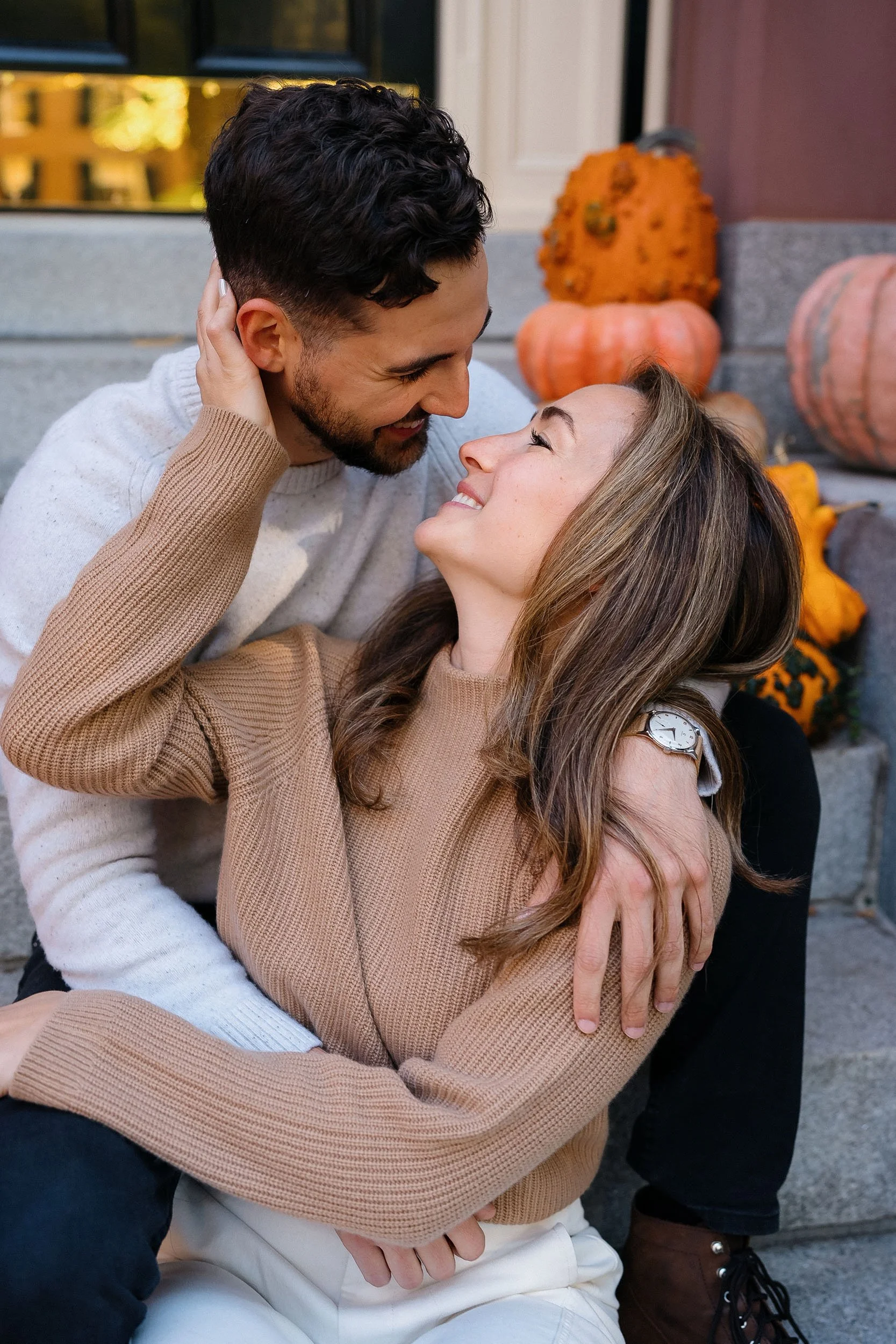 A couple sitting close together on front steps, looking at each other affectionately, with pumpkins and autumn decor in the background.