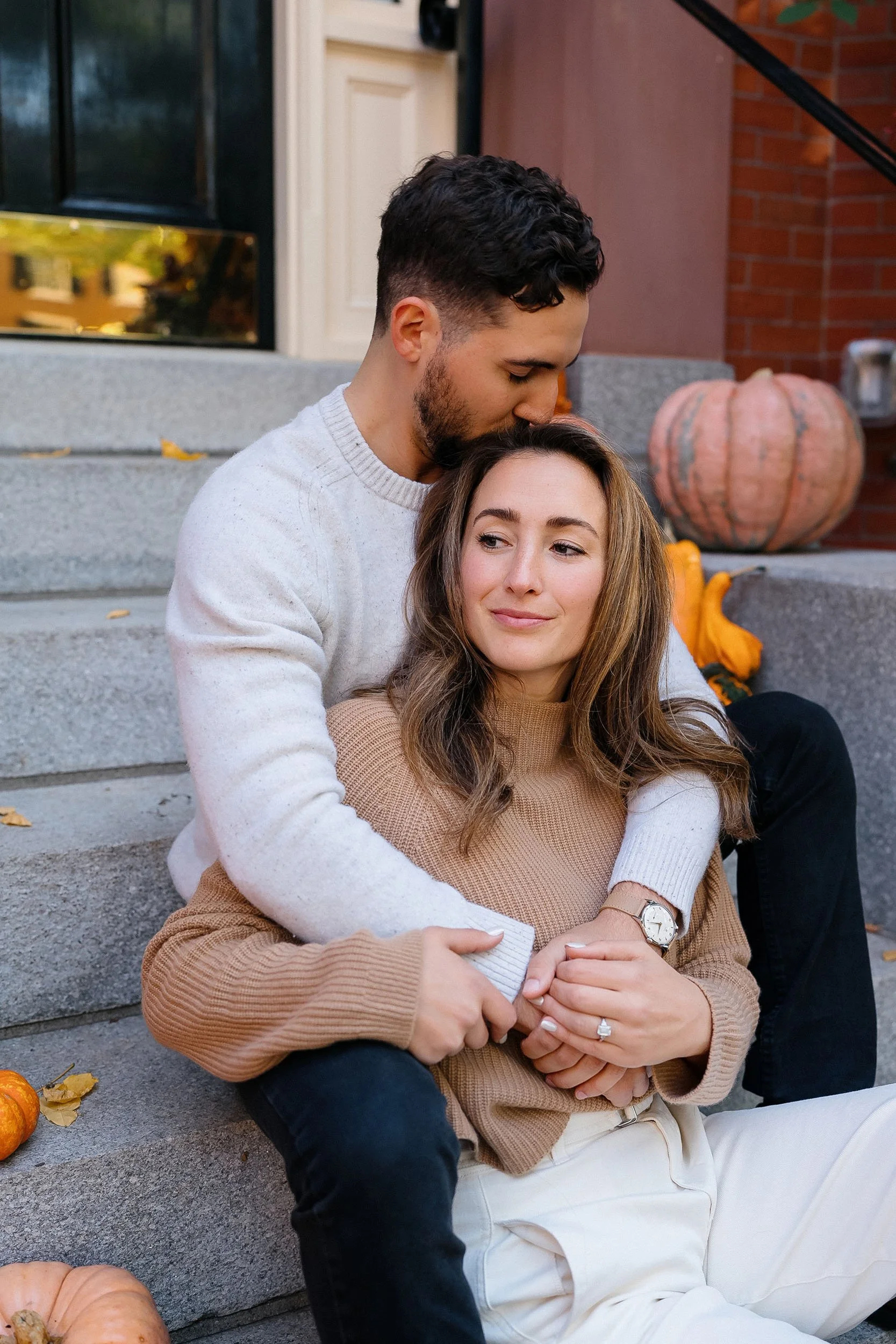 A couple sitting on front steps of a house, with pumpkins and fall leaves around them. The man is kissing the woman on the head while she looks content and relaxed.