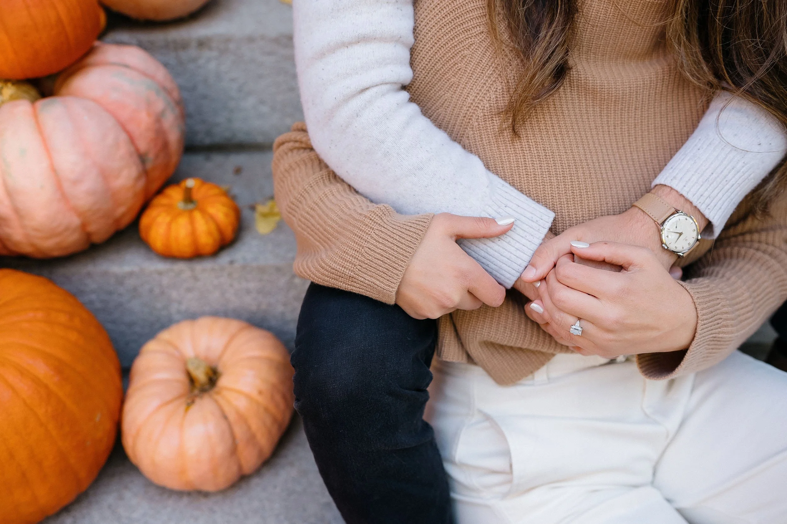 A woman sitting next to pumpkins on a wooden surface, wearing a beige and white sweater, white pants, and a watch, with her hands clasped together.