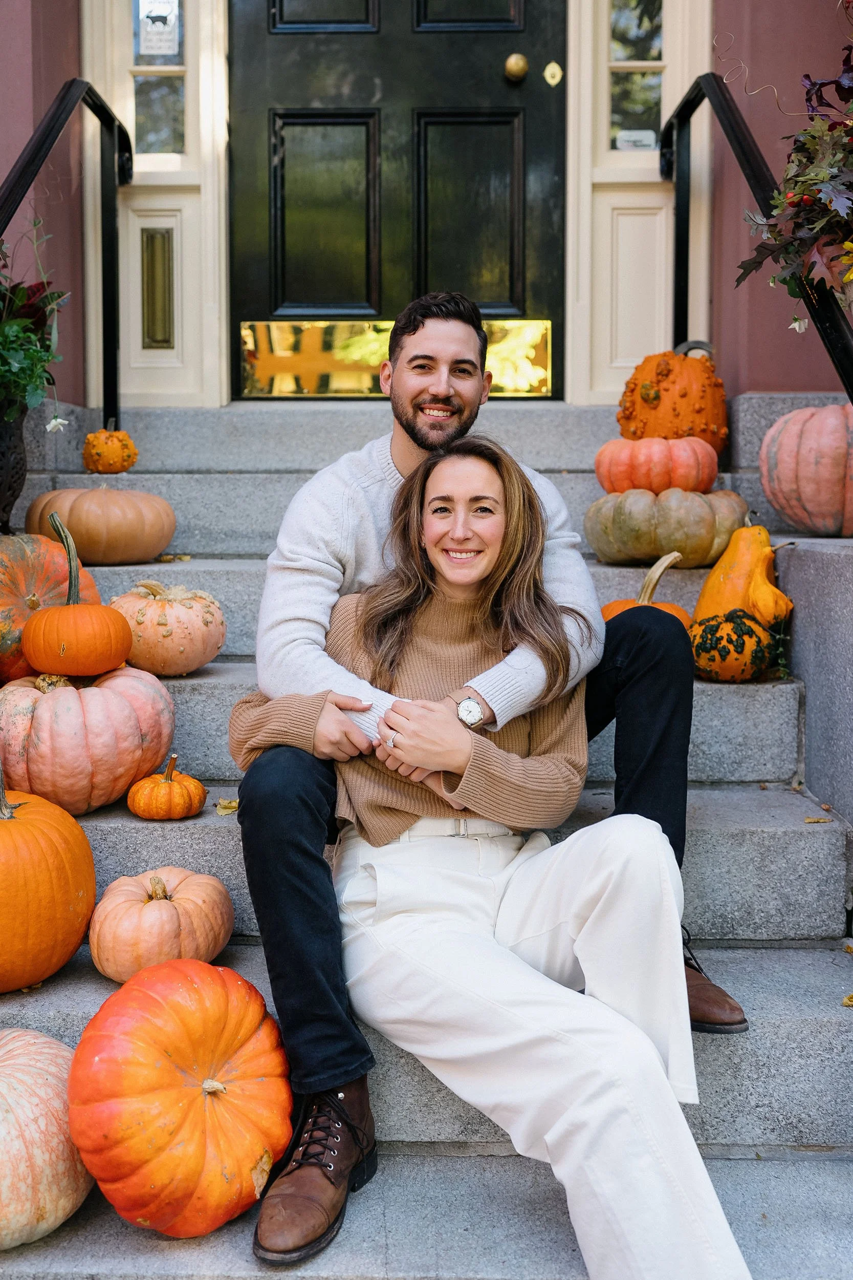 A smiling couple sitting on the front steps of their home, surrounded by pumpkins, celebrating the fall season.
