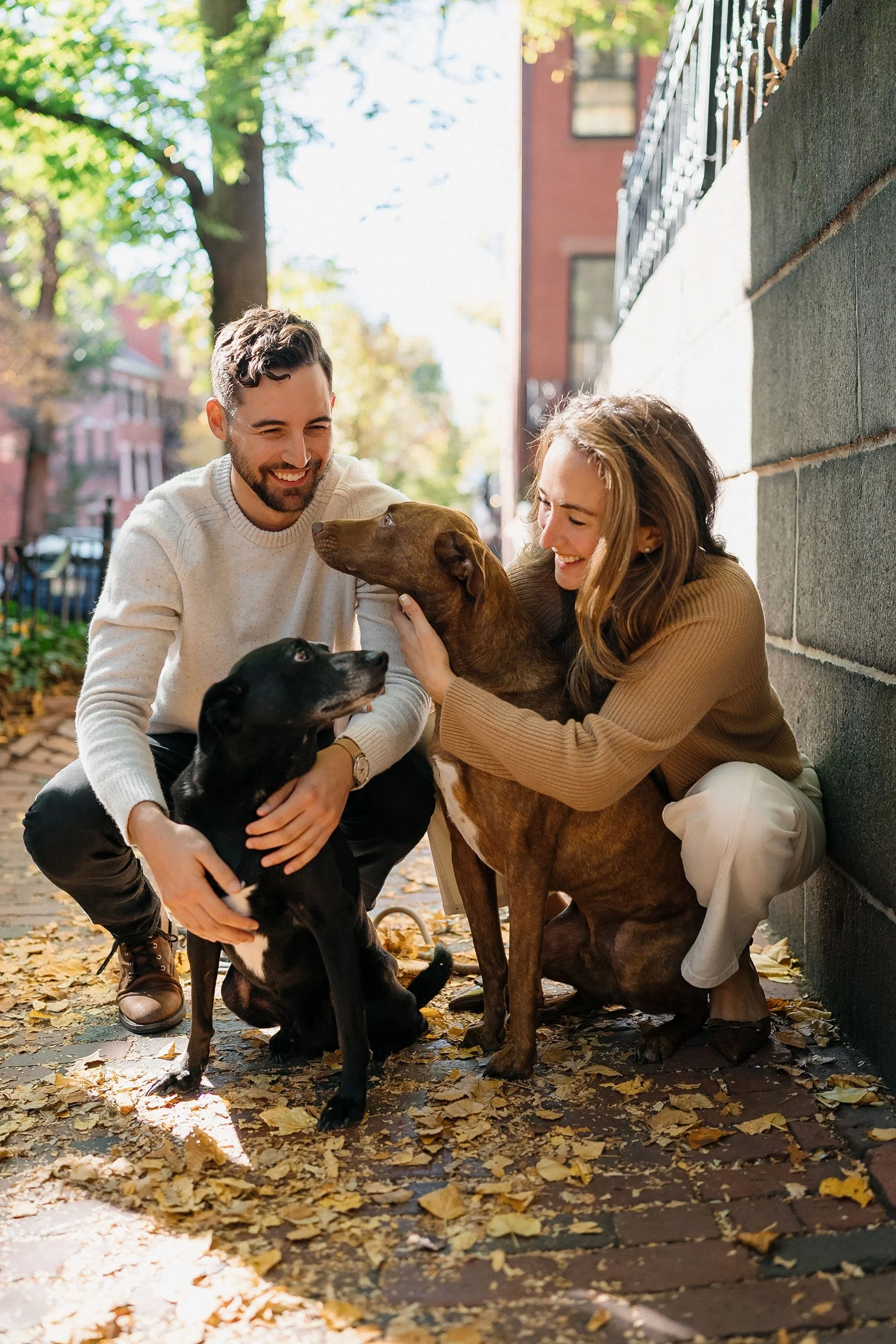 A man and woman playing with two dogs on a sidewalk covered with fallen leaves, smiling and enjoying autumn outdoors.