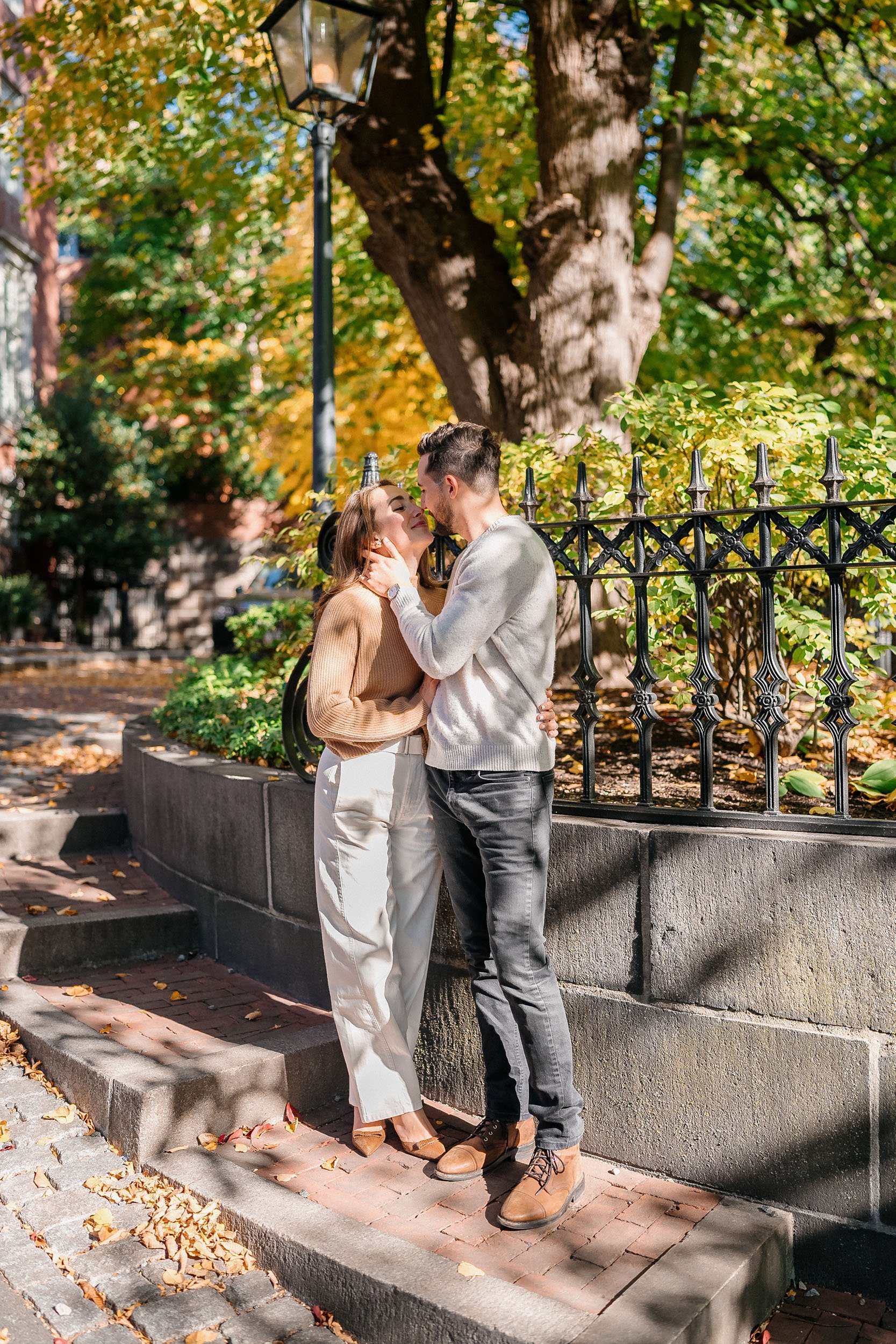A couple sharing an intimate moment on a city sidewalk with autumnal trees and foliage in the background.