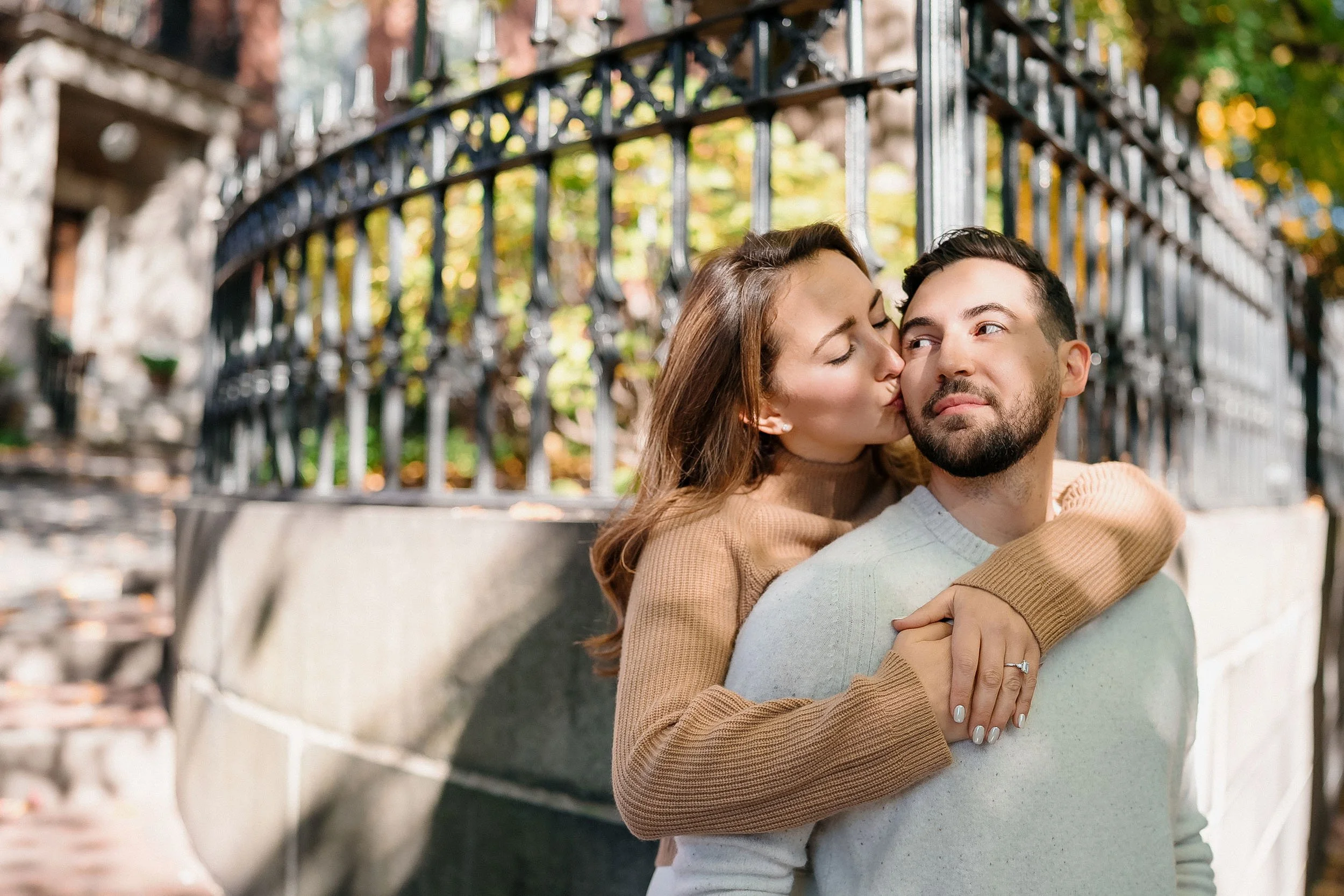 A woman with brown hair and a tan sweater kisses a man with beard and a light gray sweater on the cheek outdoors in front of a black iron fence, with trees and sunlight in the background.