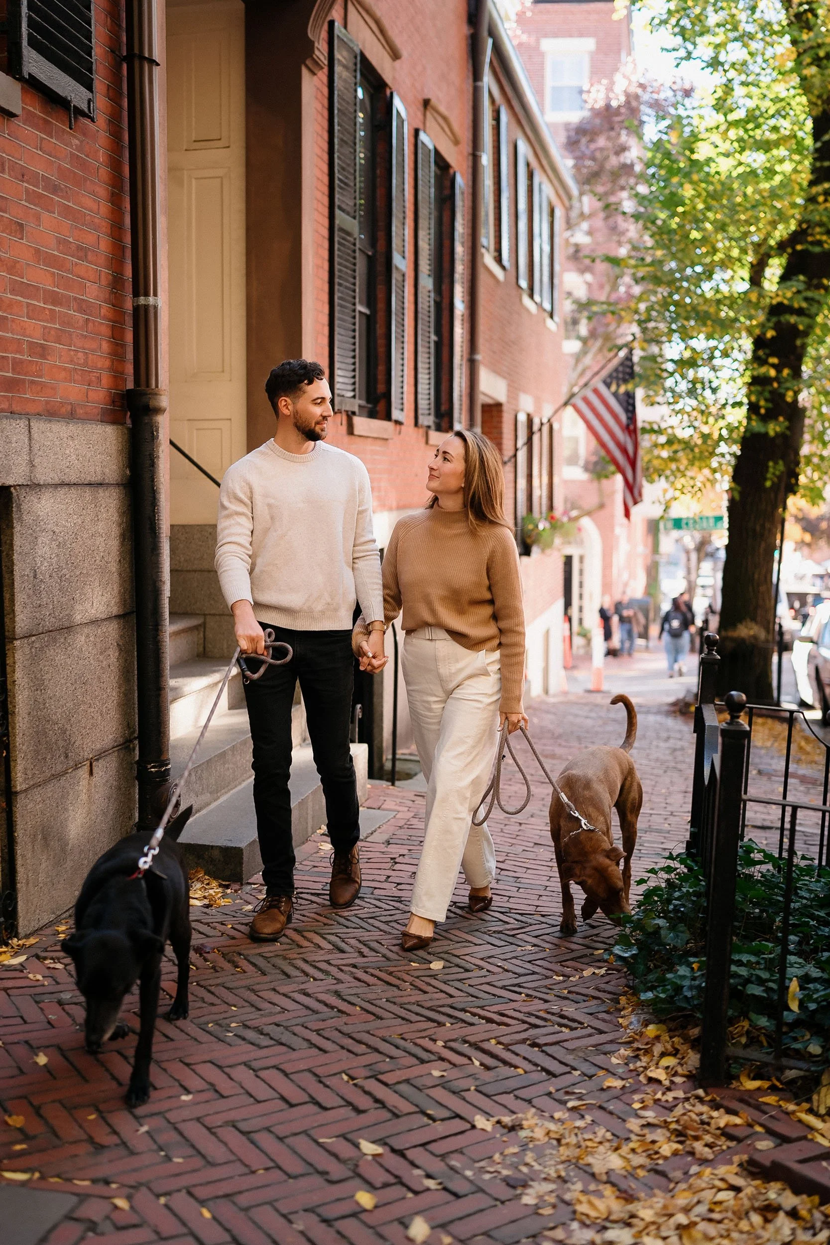 A young couple walks hand in hand with two dogs on a brick sidewalk in a neighborhood with red brick townhouses and trees. The man has a beard and wears a cream sweater and black pants. The woman has long hair and wears a brown sweater and white pant