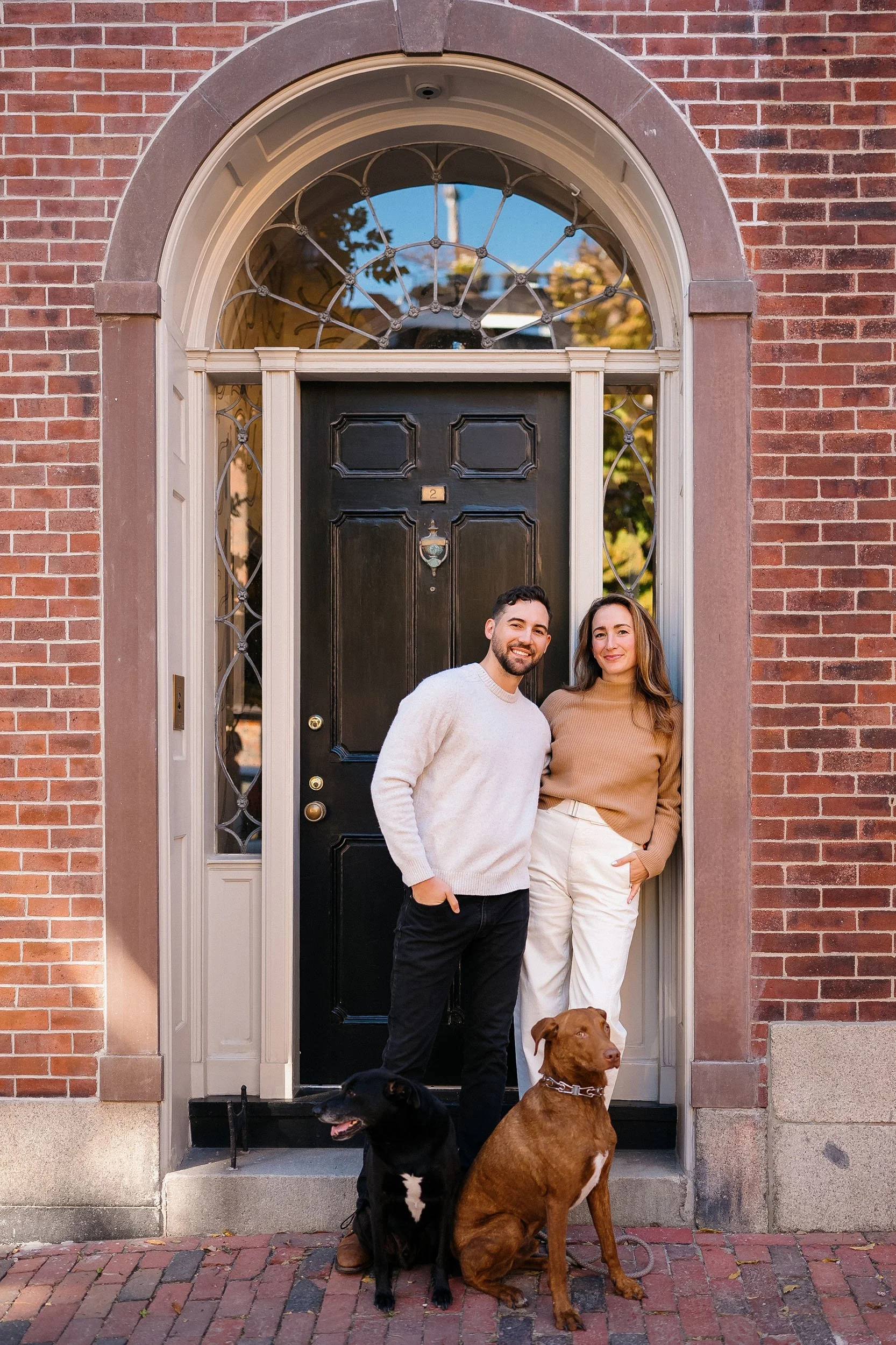 A couple standing by their front door with two dogs, in front of a brick house with a black door and decorative glass windows.