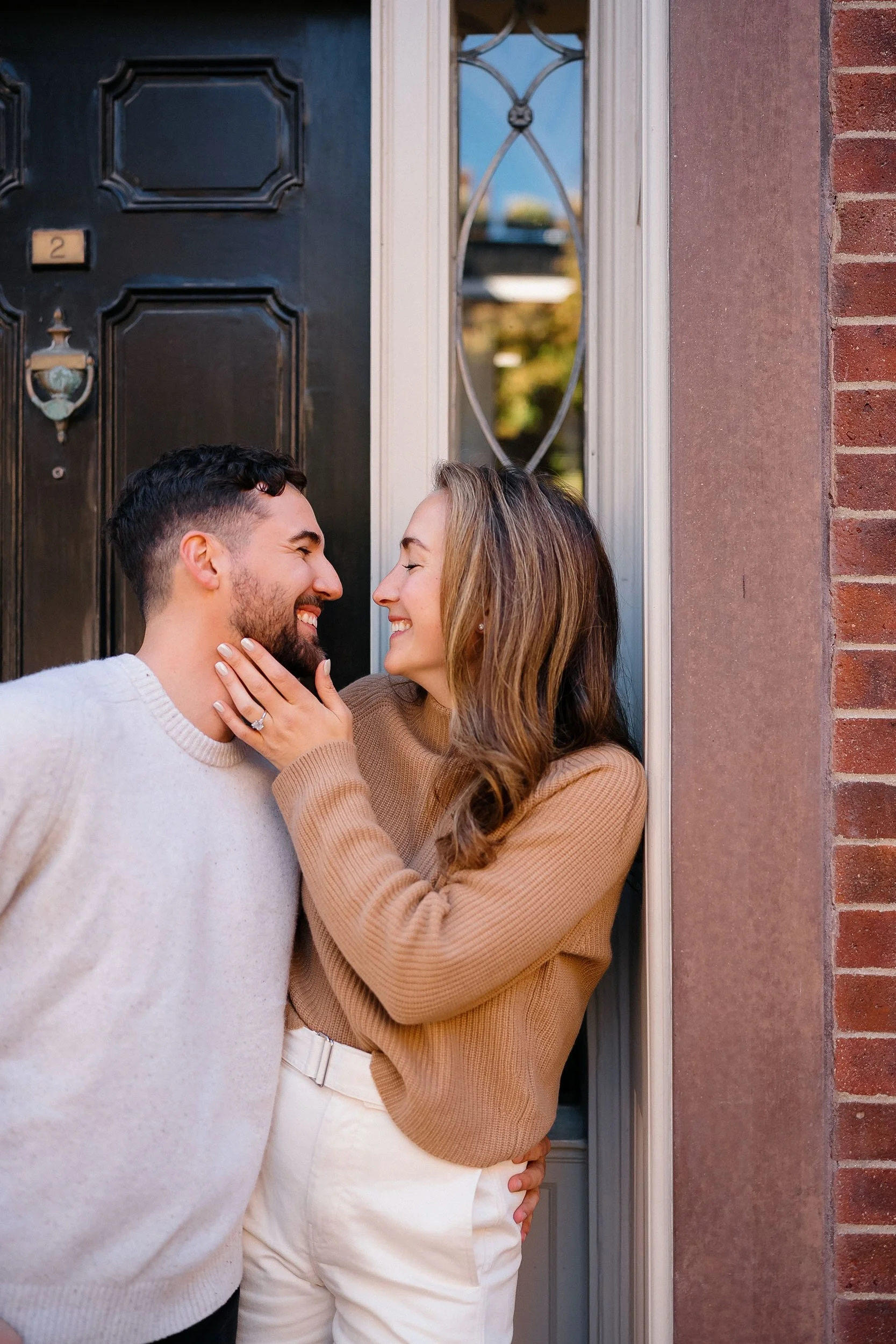 A couple smiling and looking at each other affectionately outside a front door. The woman is touching the man's face and wearing an engagement ring.