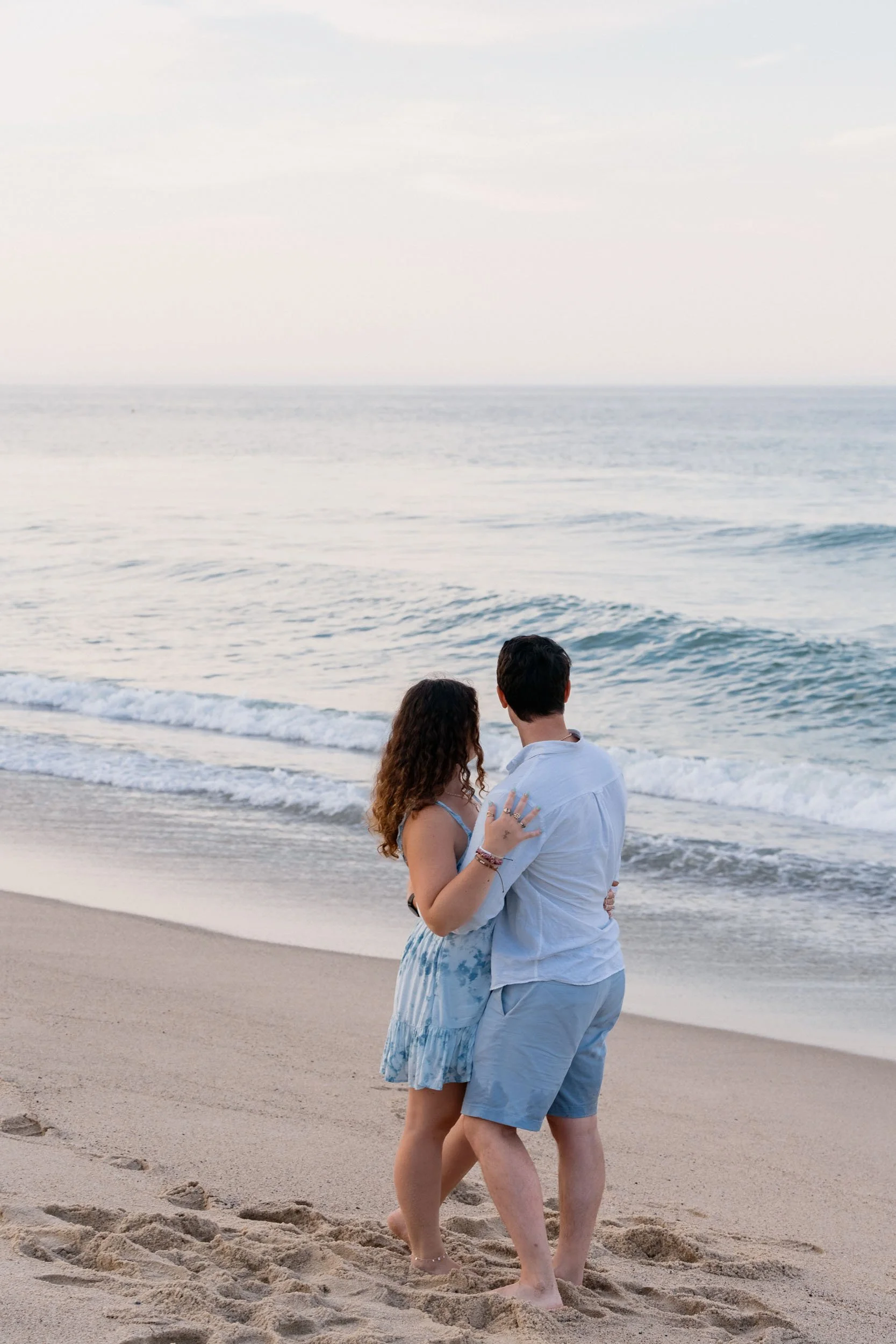 A couple standing close together on a sandy beach, looking at the ocean waves during sunset.