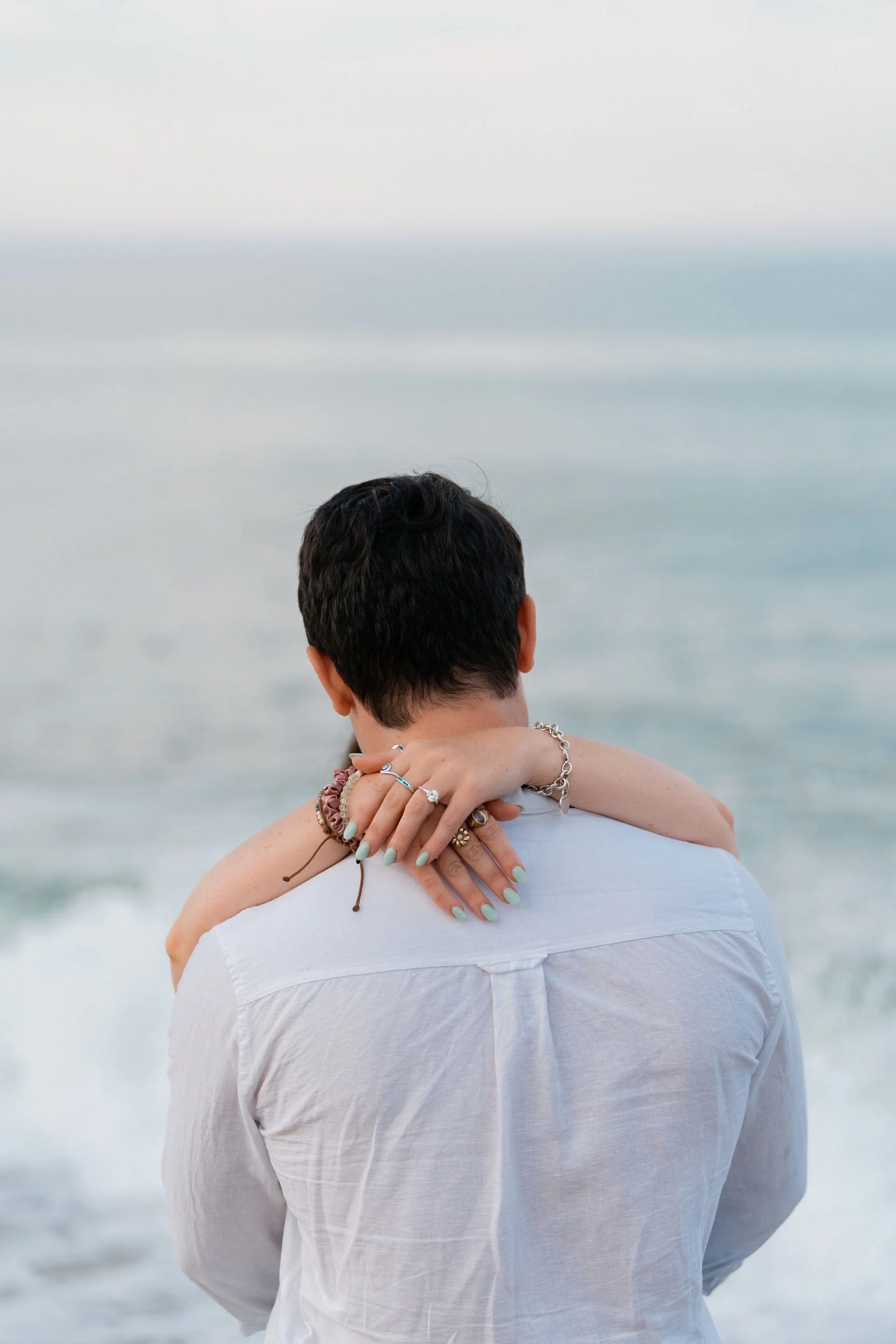 A person with short dark hair faces away, wearing a white shirt, while another person's arms with jewelry hug them from behind at the beach with ocean waves in the background.