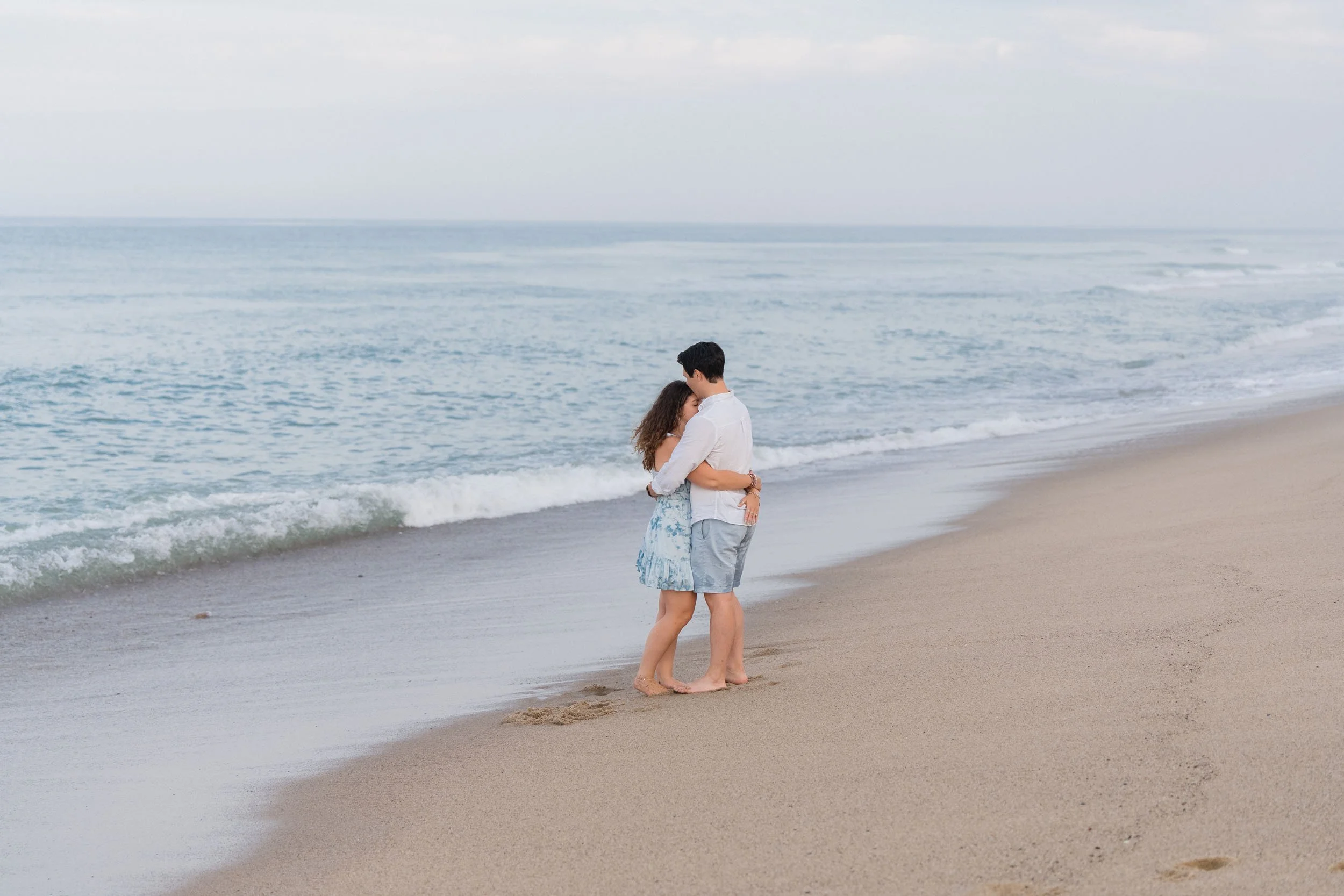 A young couple hugging on a sandy beach near the ocean, with the waves in the background.