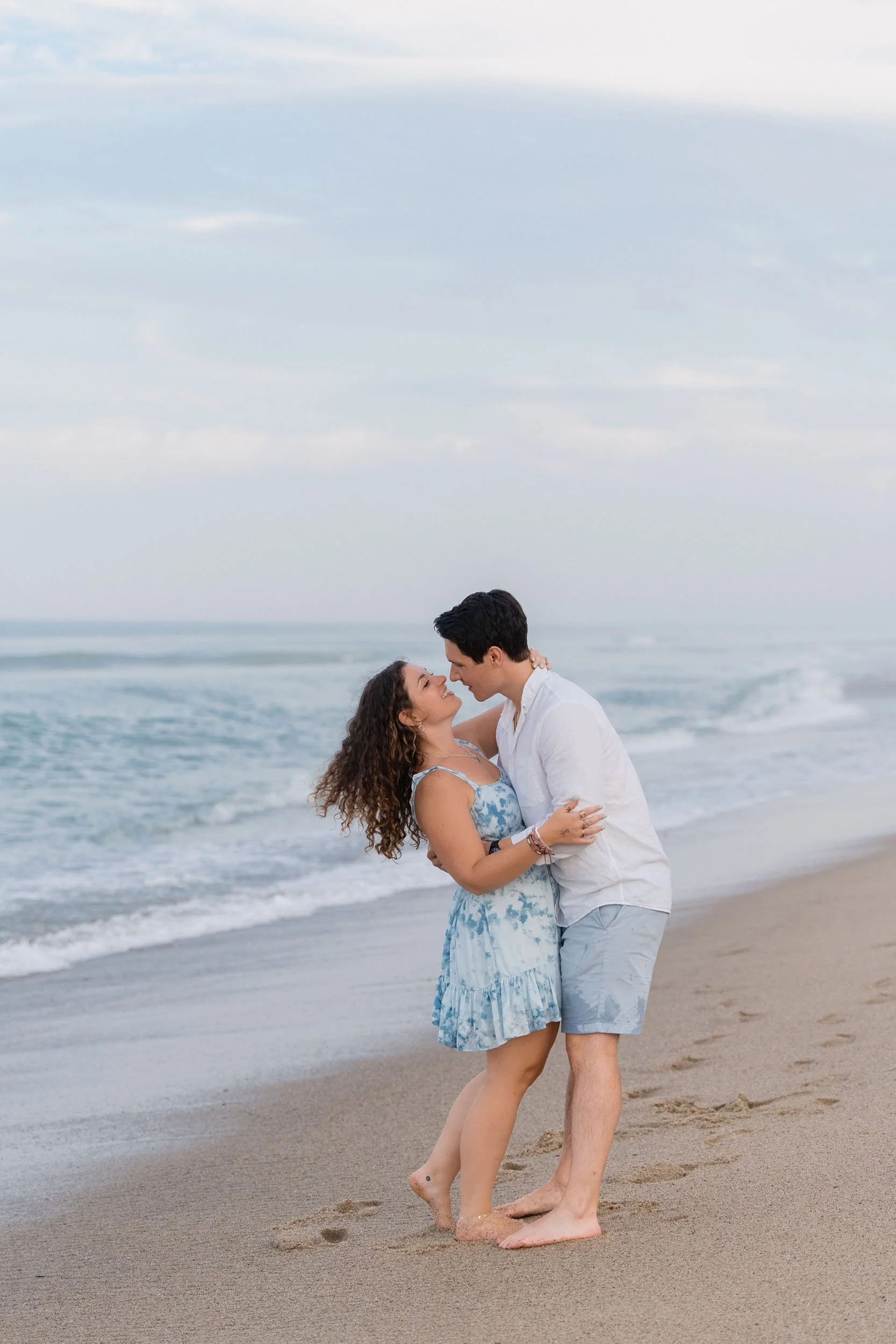 A couple embracing and about to kiss on the beach with the ocean and sky in the background.