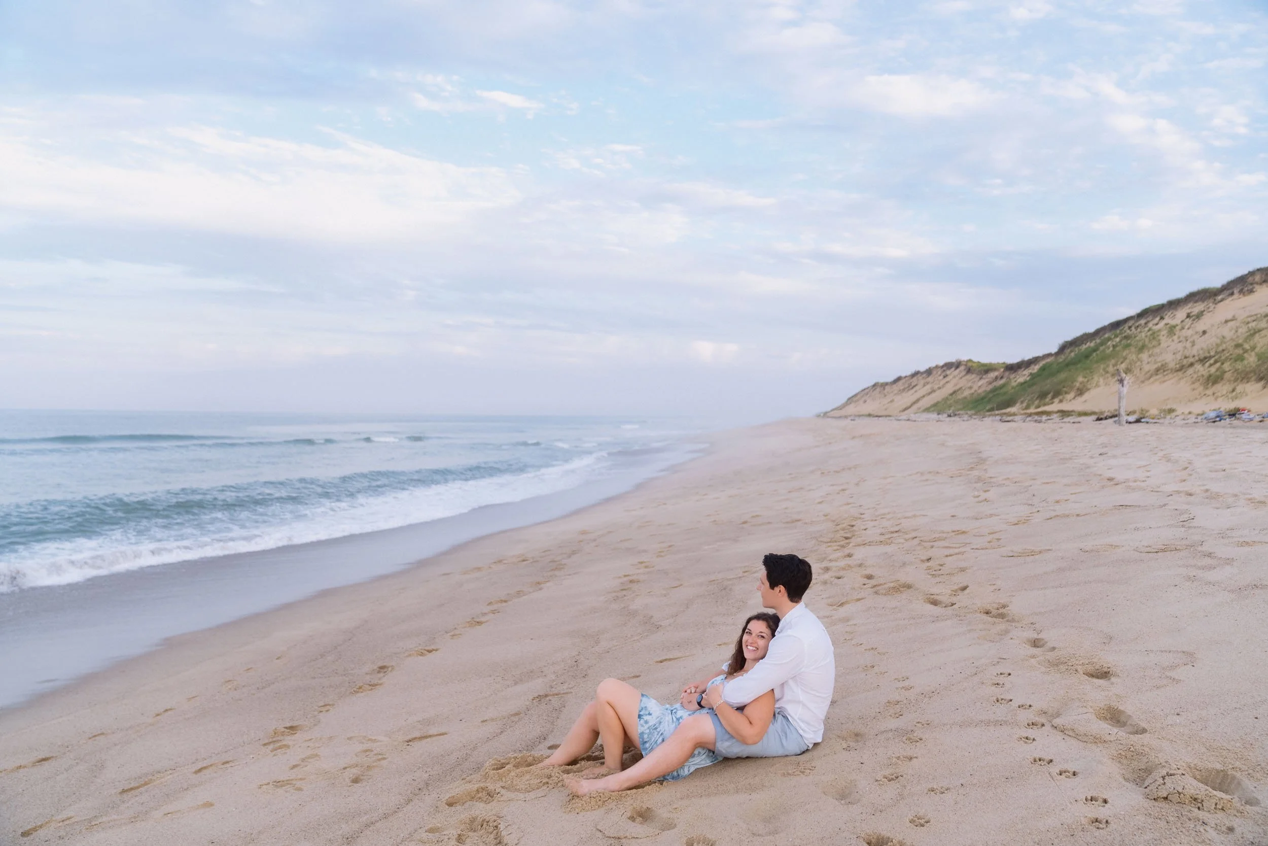 A couple sits on a sandy beach, facing the ocean, smiling and enjoying the scenery with dunes and a cloudy sky in the background.