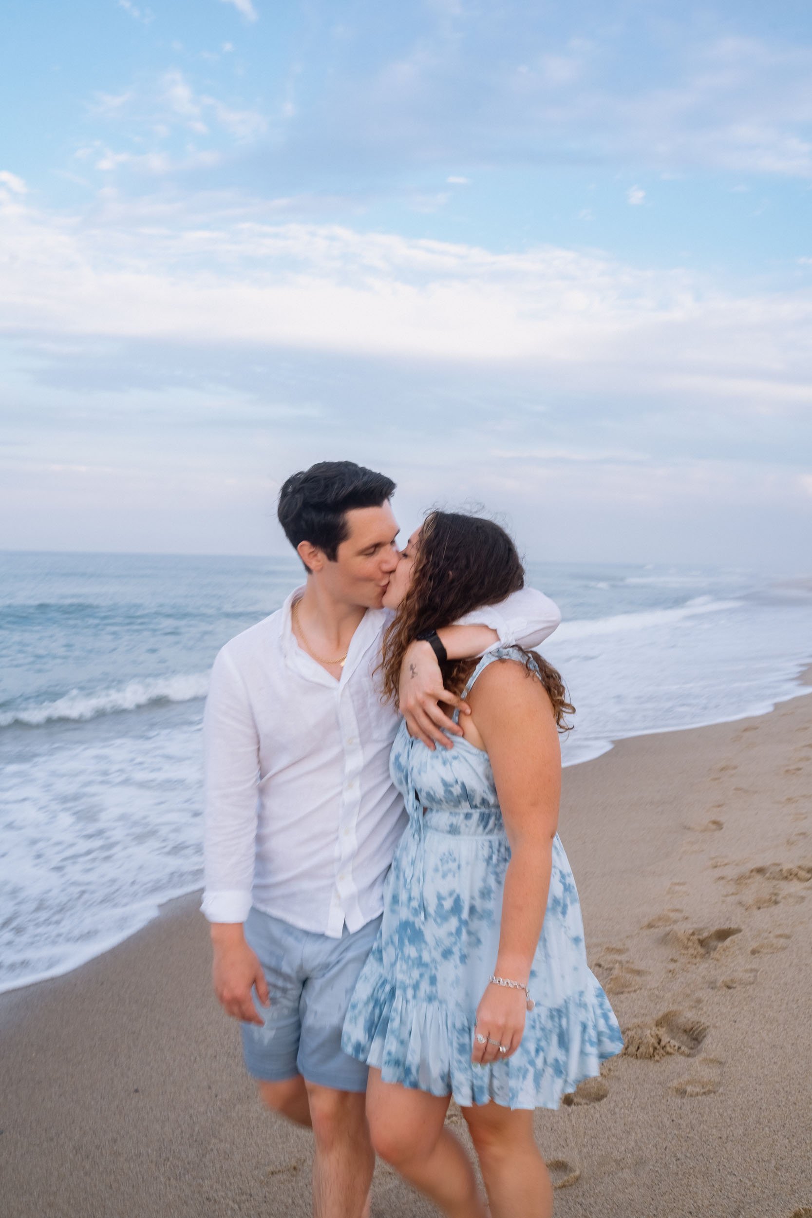 A young couple sharing a kiss on the beach near the ocean, with footprints in the sand and a partly cloudy sky overhead.