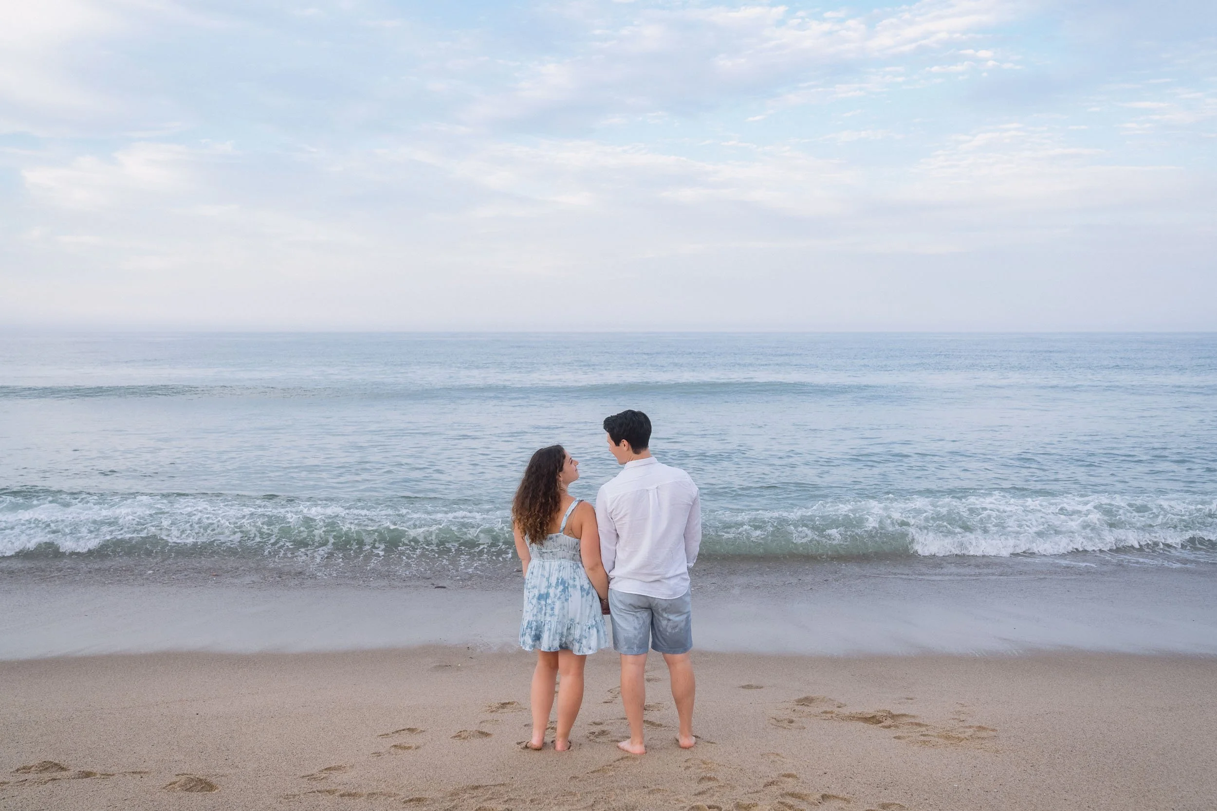 A young couple stands hand-in-hand on the sandy beach, facing the ocean, with gentle waves and a cloudy sky above.