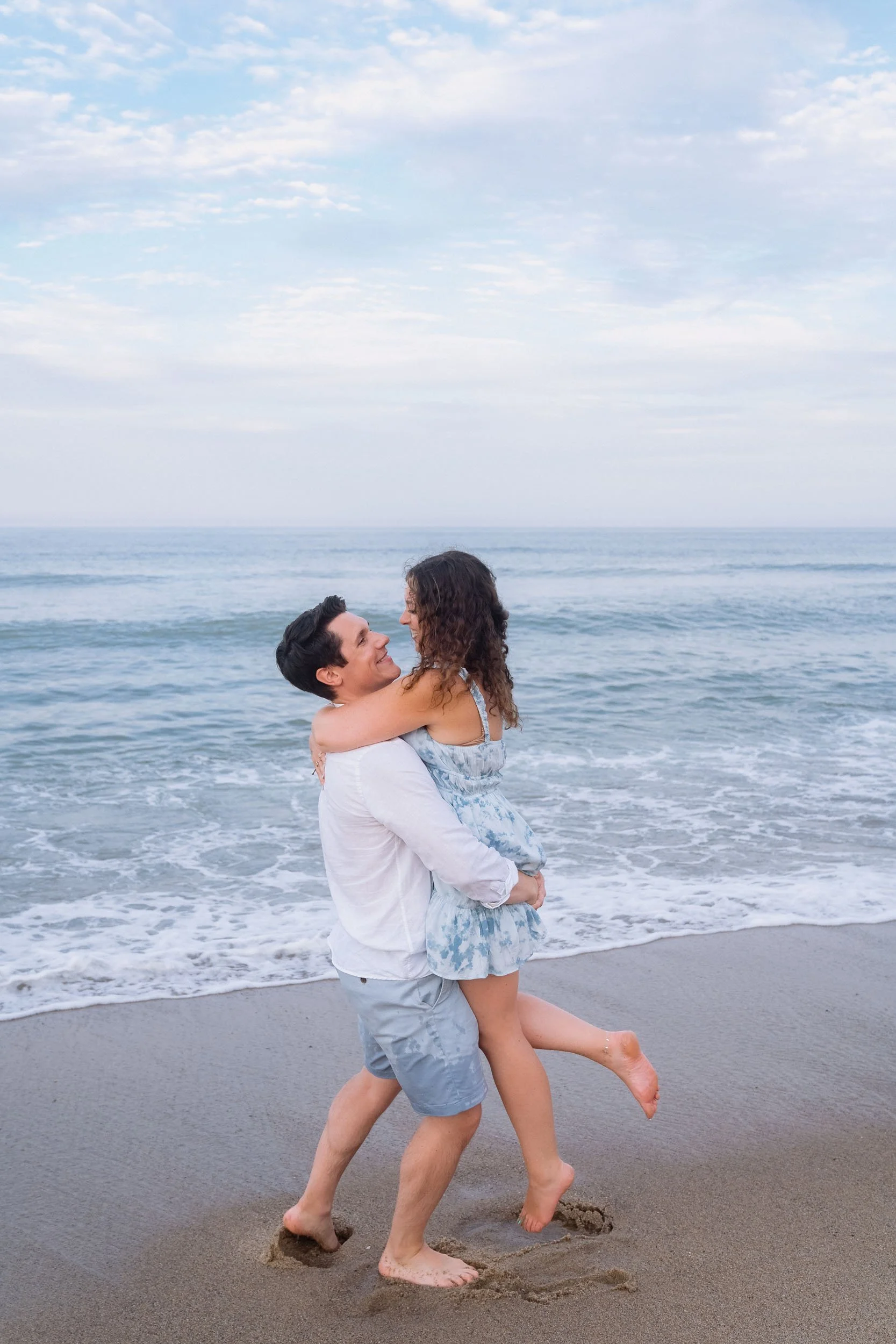 A couple on the beach, the man lifting the woman in his arms while standing on the sand near the ocean waves, with a cloudy sky overhead.