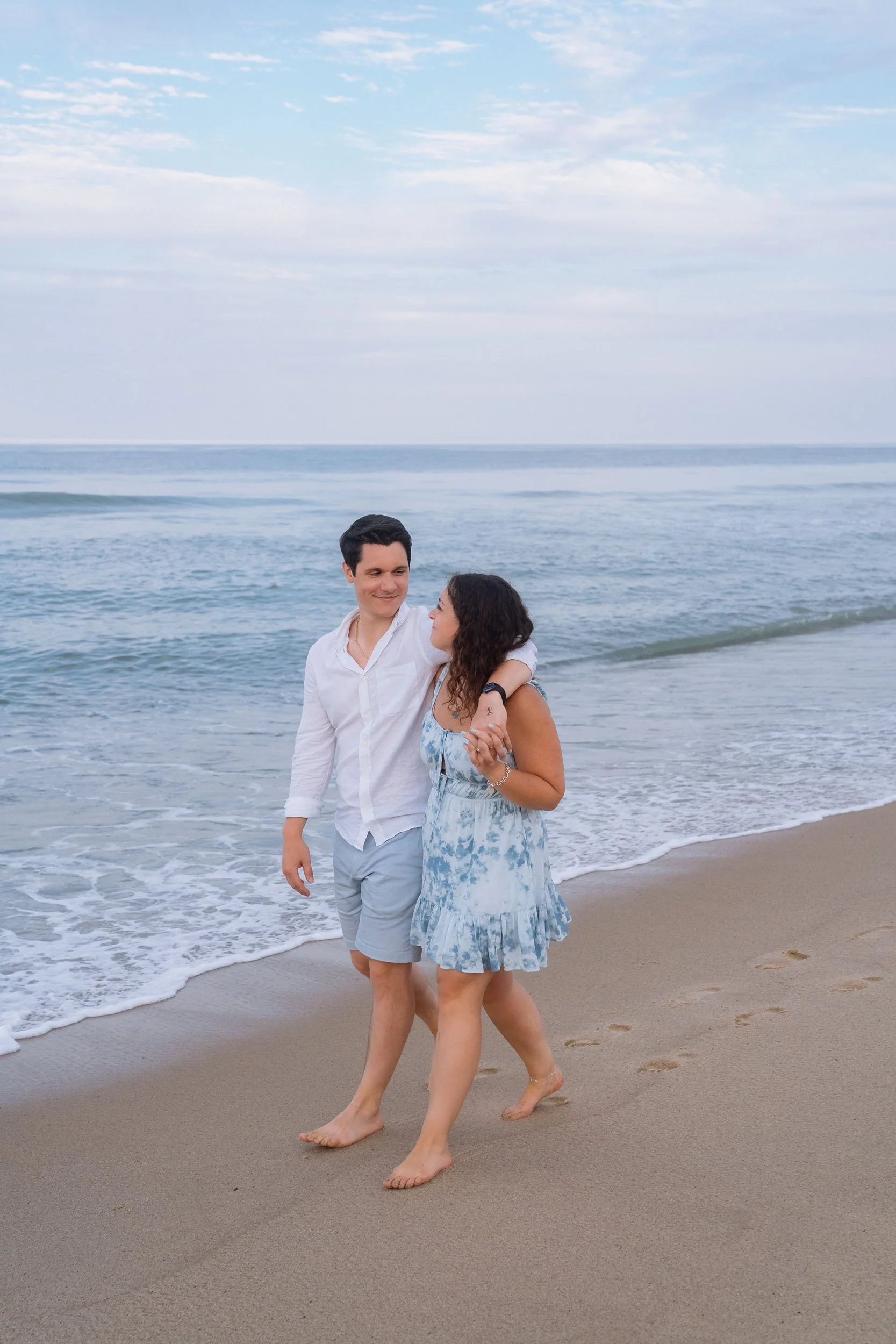 A young couple walking along the beach, holding hands and looking at each other, with ocean waves and a cloudy sky in the background.
