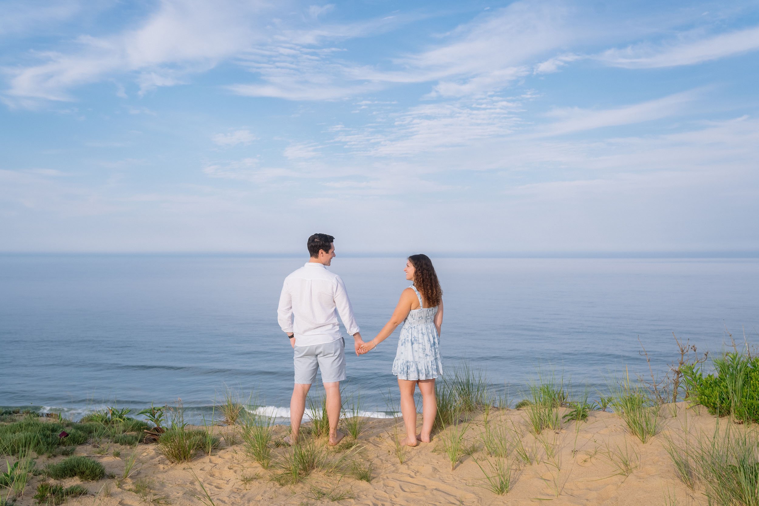A couple holding hands on a sandy beach, facing the ocean with a blue sky and clouds above.