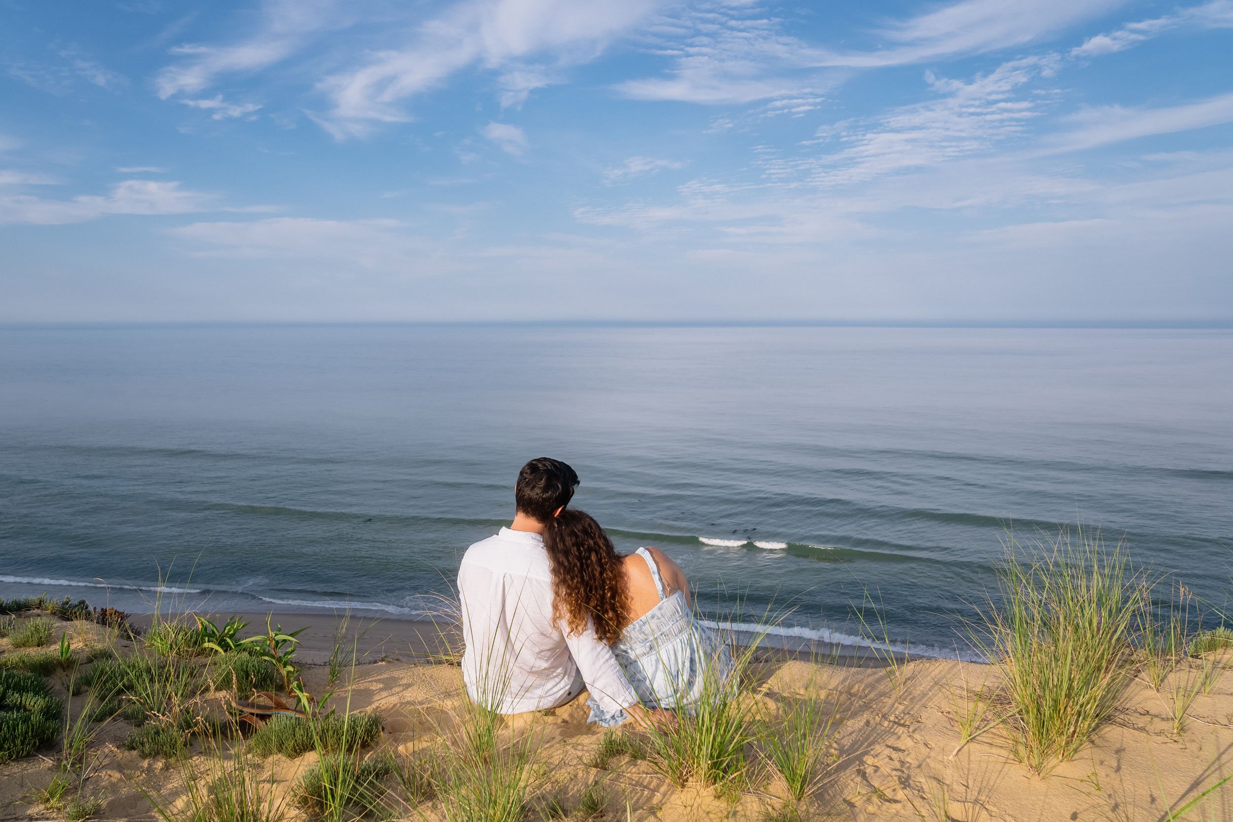 A couple sitting on a sandy beach with grass, looking out at the calm ocean under a partly cloudy sky.