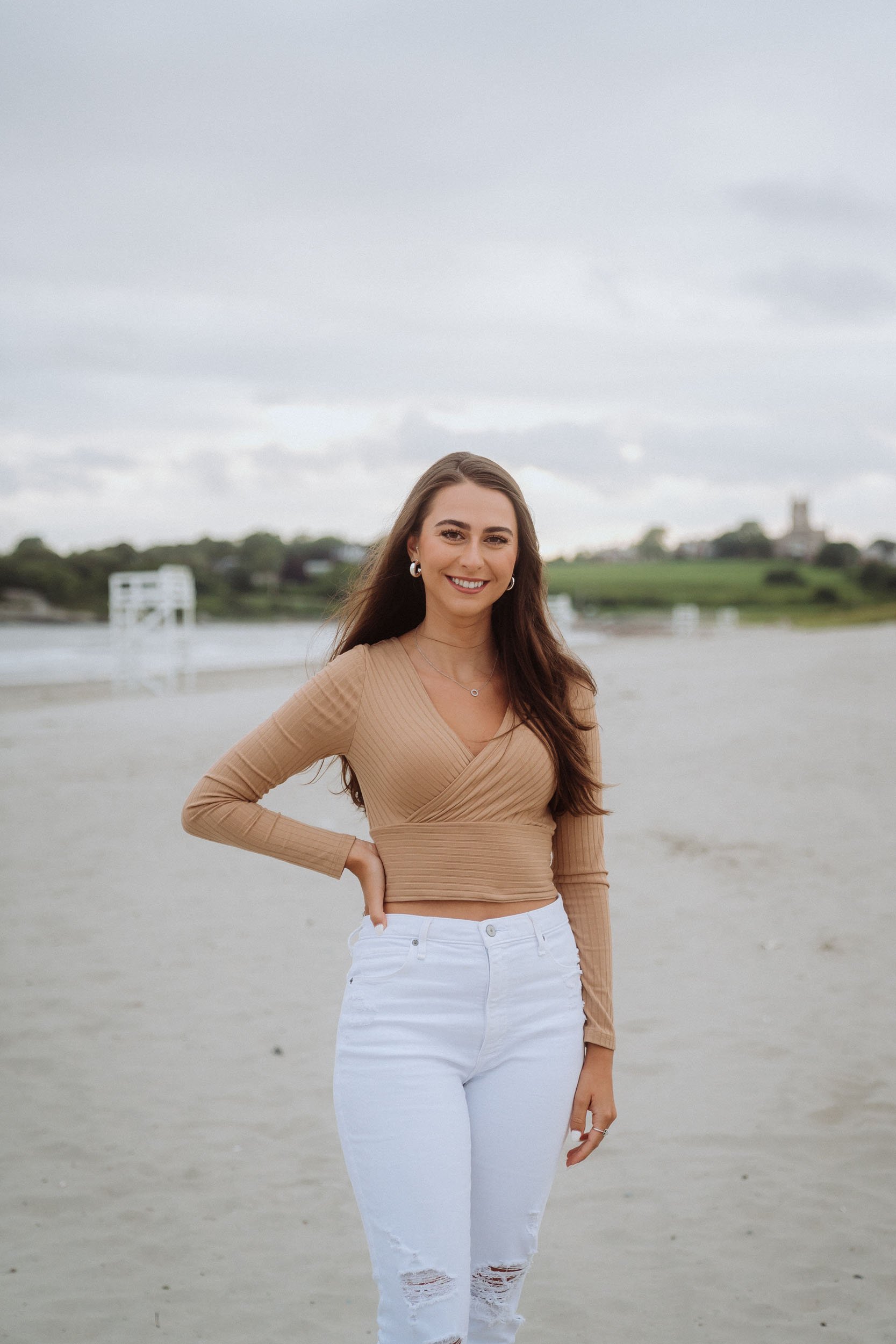 A woman with long brown hair and a bright smile standing on a sandy beach with water and greenery in the background, wearing a tan long-sleeve top and white ripped jeans.