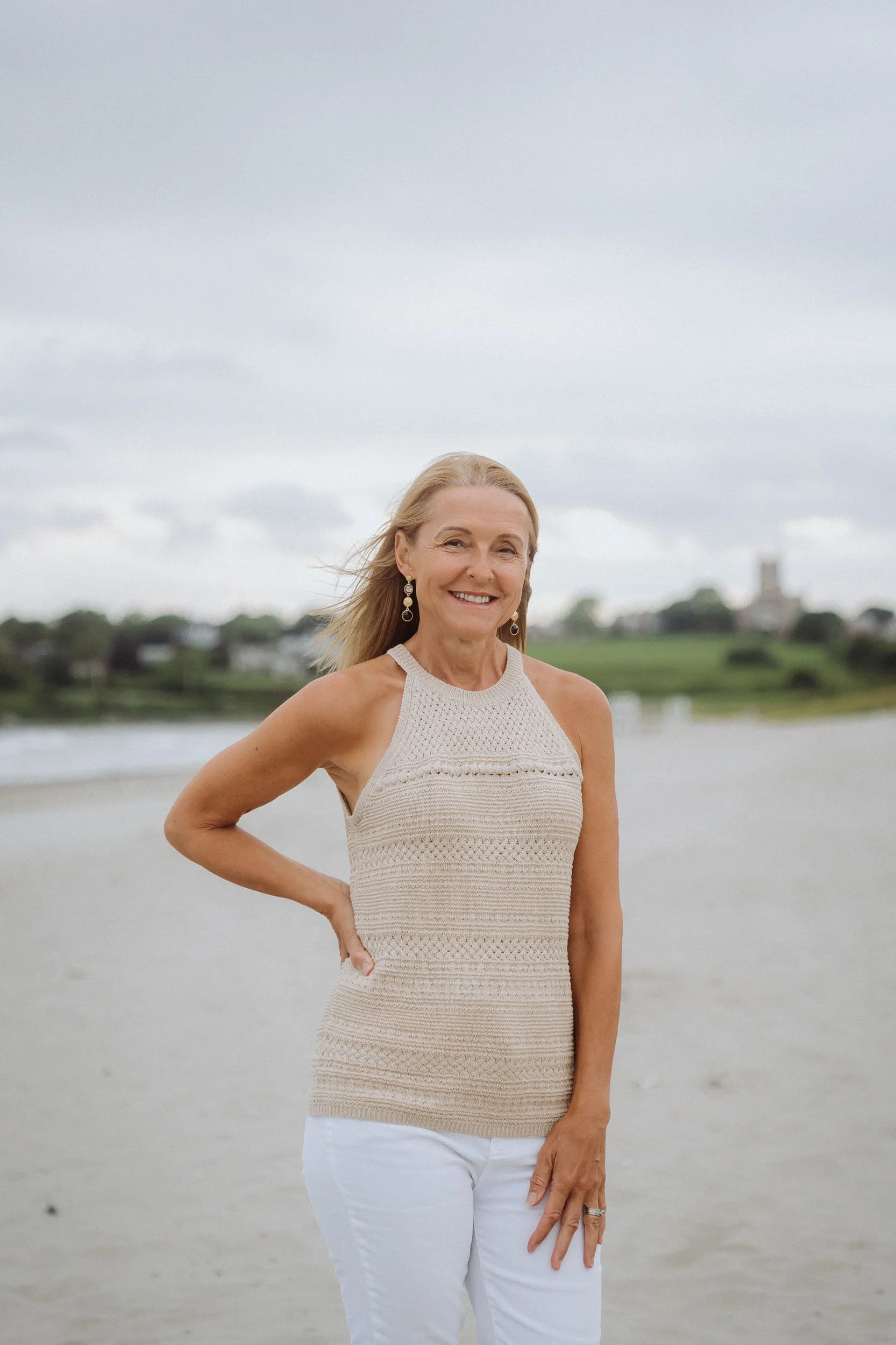 Smiling middle-aged woman with blonde hair in a sleeveless beige knit top and white pants standing on a beach with water and cloudy sky in the background.