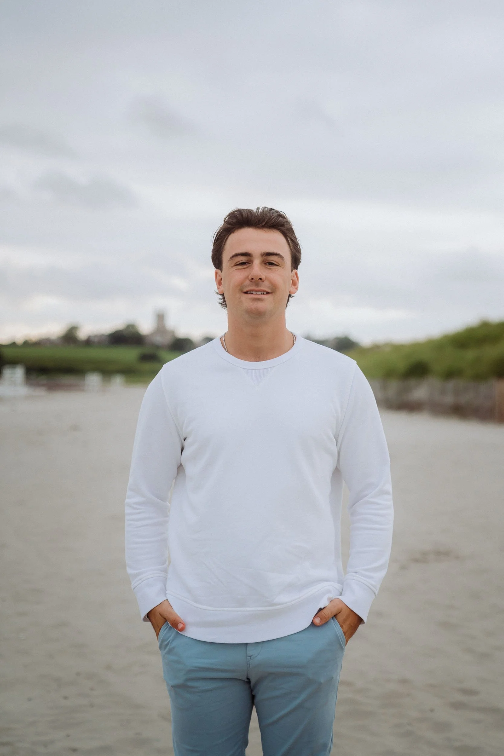 Young man standing on a sandy beach with a cloudy sky, green hills, and a distant building or castle in the background.