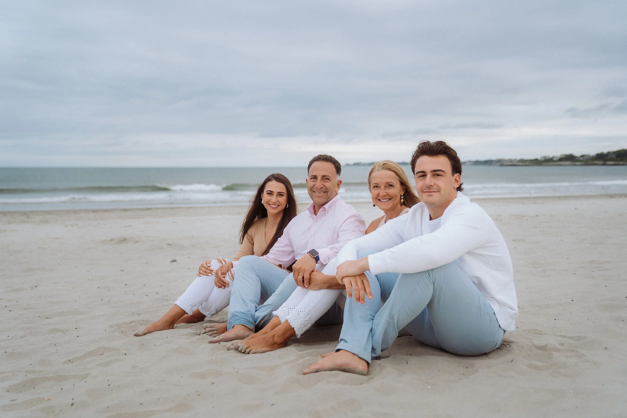 Four people sitting on the beach sand, smiling, with the ocean and cloudy sky in the background.