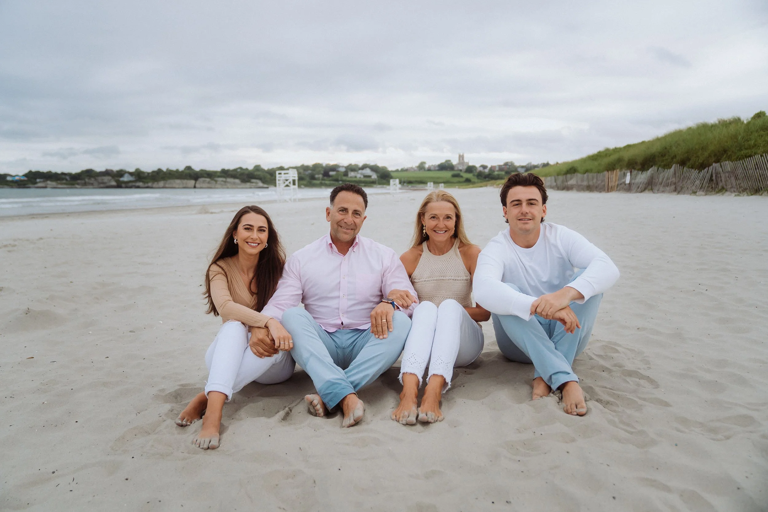 Four people sitting on a sandy beach, smiling at the camera, with ocean, sand dunes, and cloudy sky in the background.
