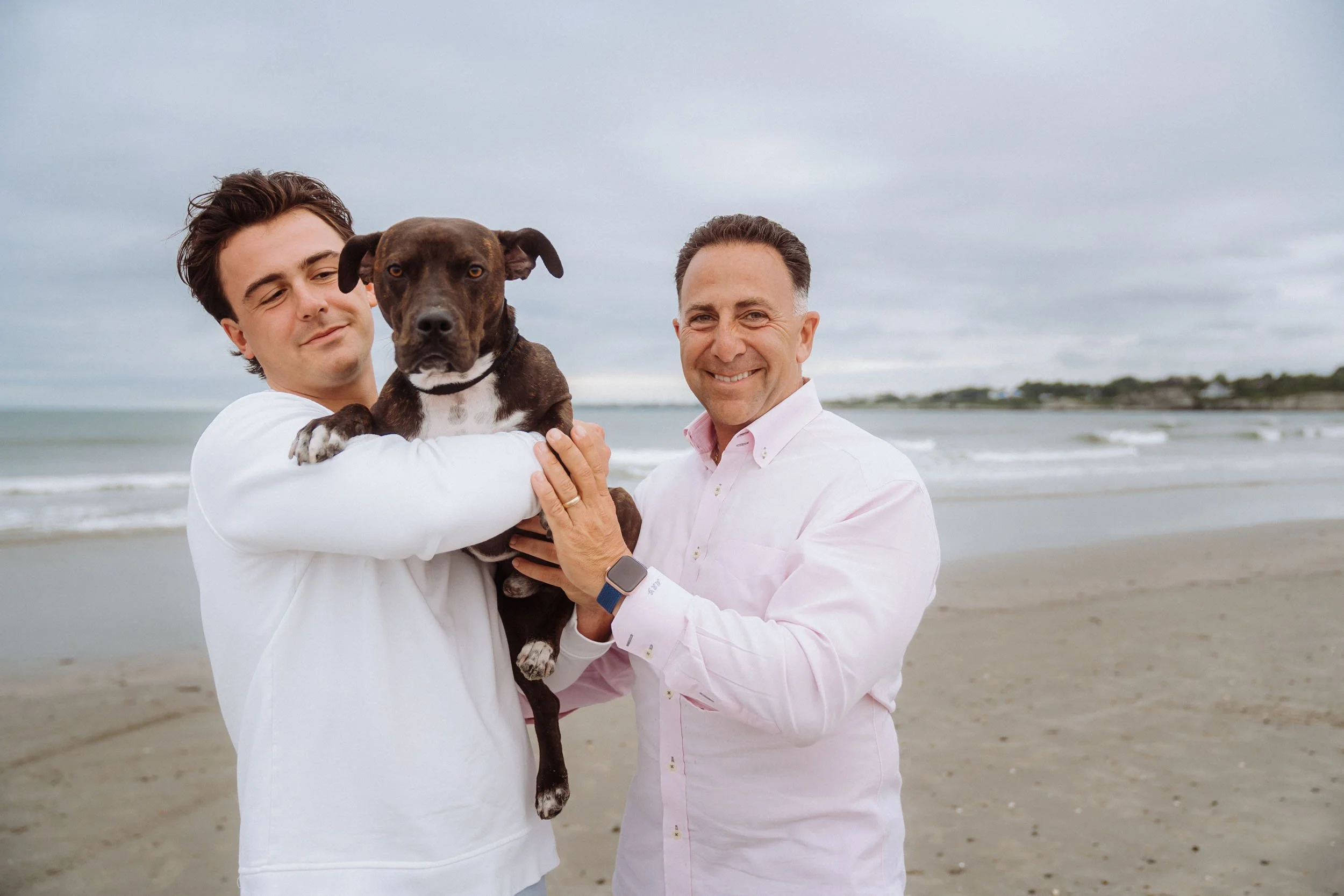 Two men standing on a beach holding a brown and white dog, with an overcast sky and ocean in the background.