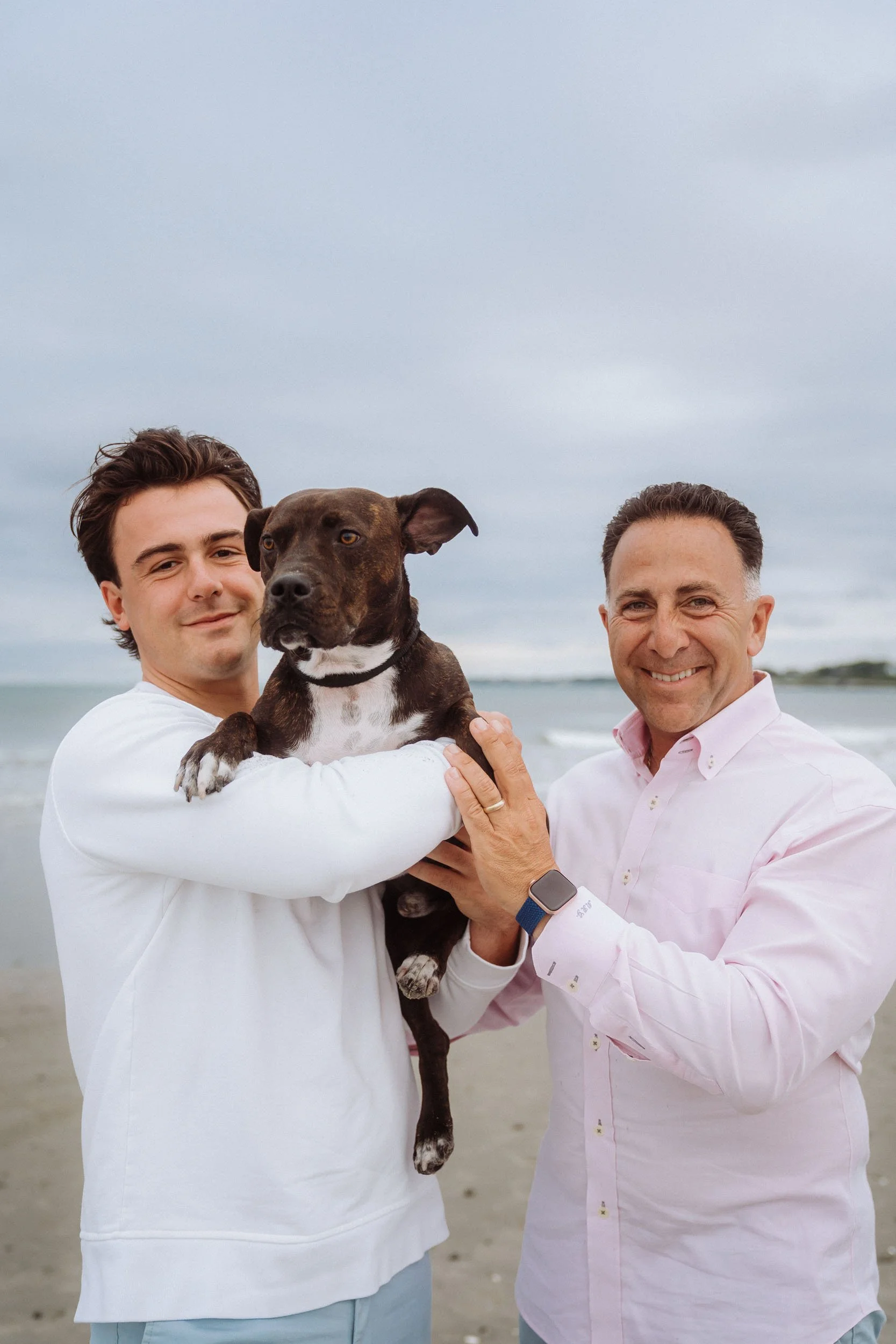 Two men holding a brown and white dog on a beach, with ocean waves and overcast sky in the background.